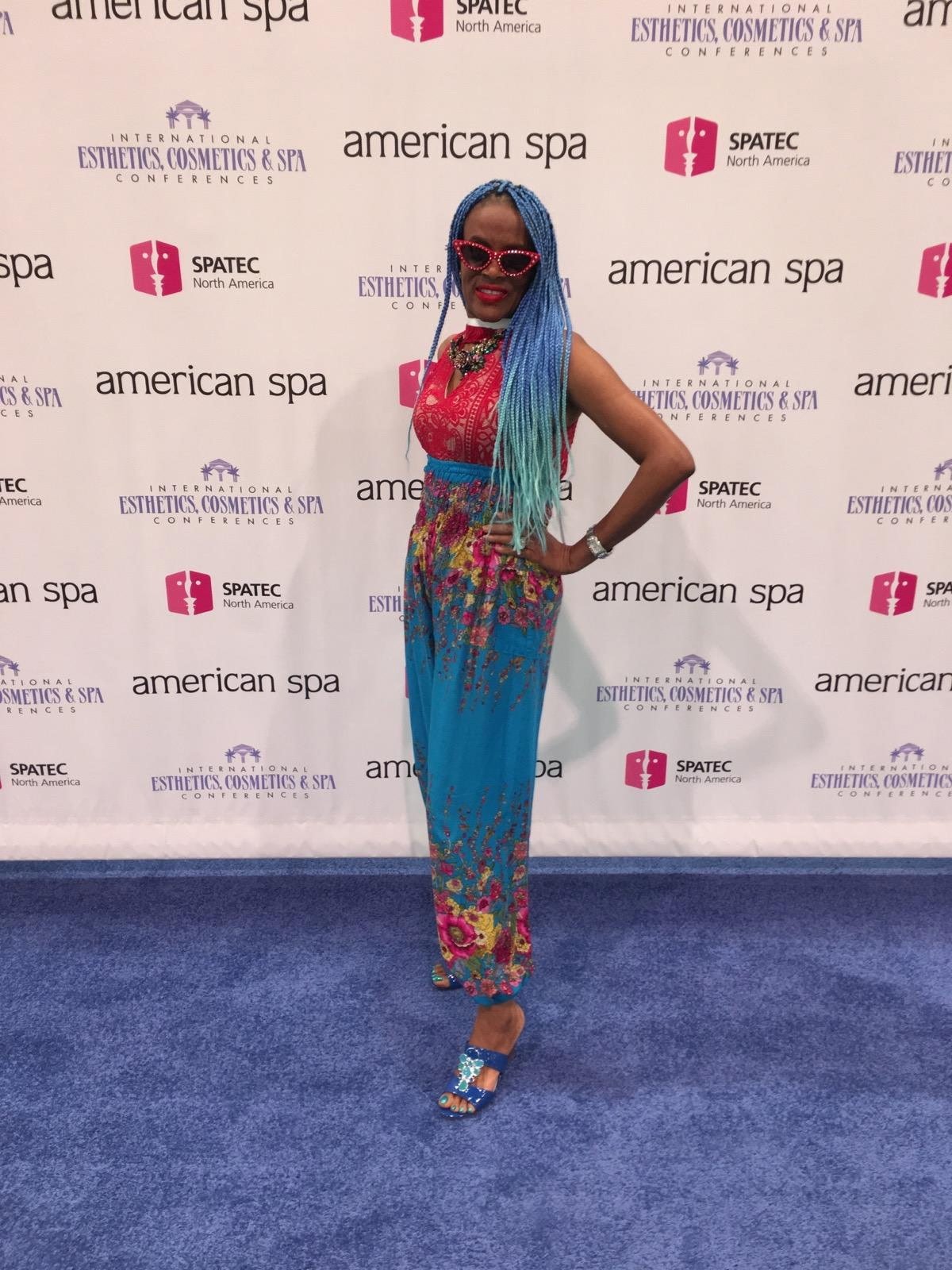 Person with long blue and turquoise braided hair and red sunglasses posing in front of a backdrop with logos for International Esthetics, Cosmetics & Spa Conferences and American Spa.