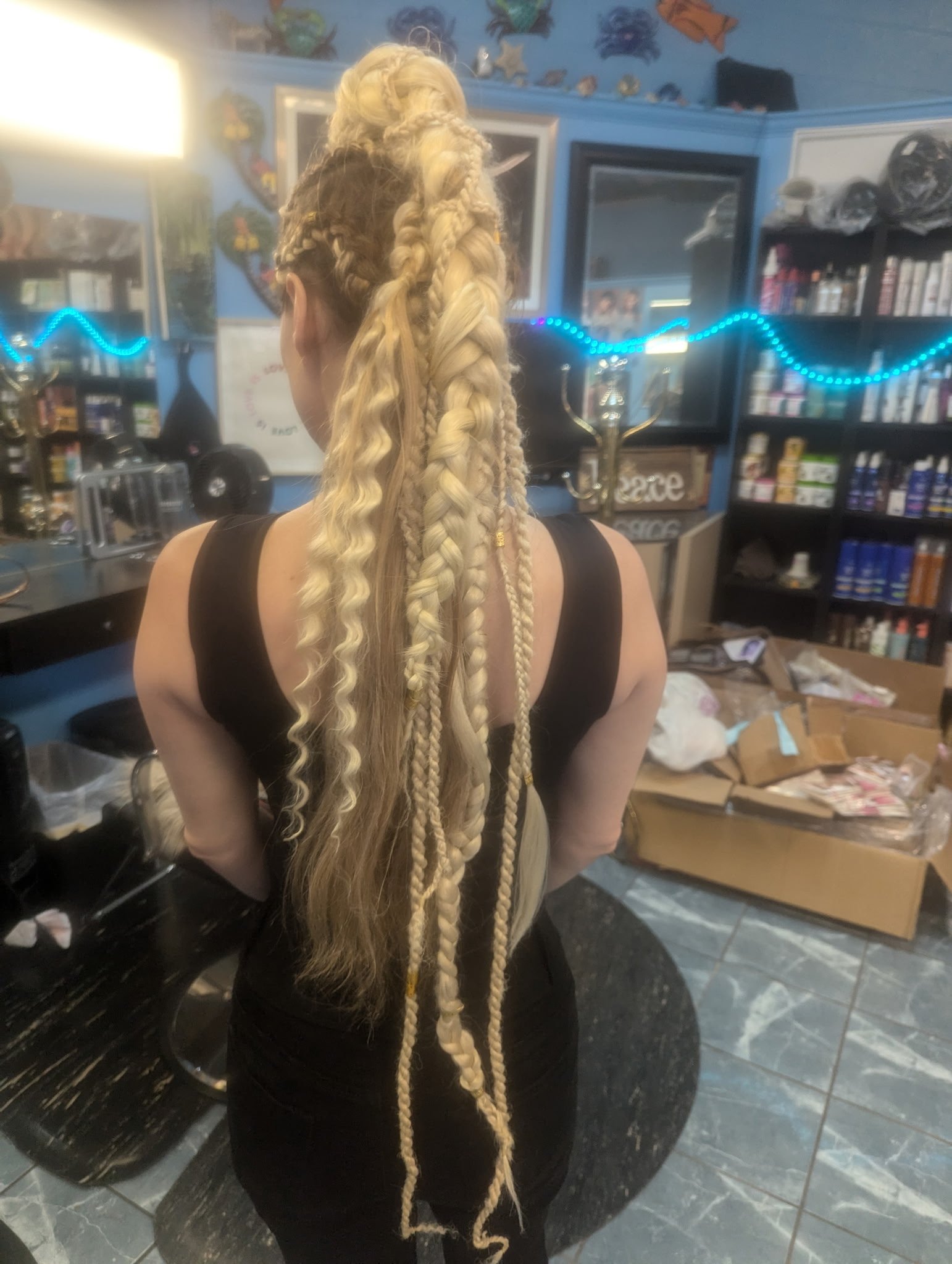A woman with long blonde braided hair, featuring intricate fishtail and rope braids, standing in a salon with various hair products in the background.