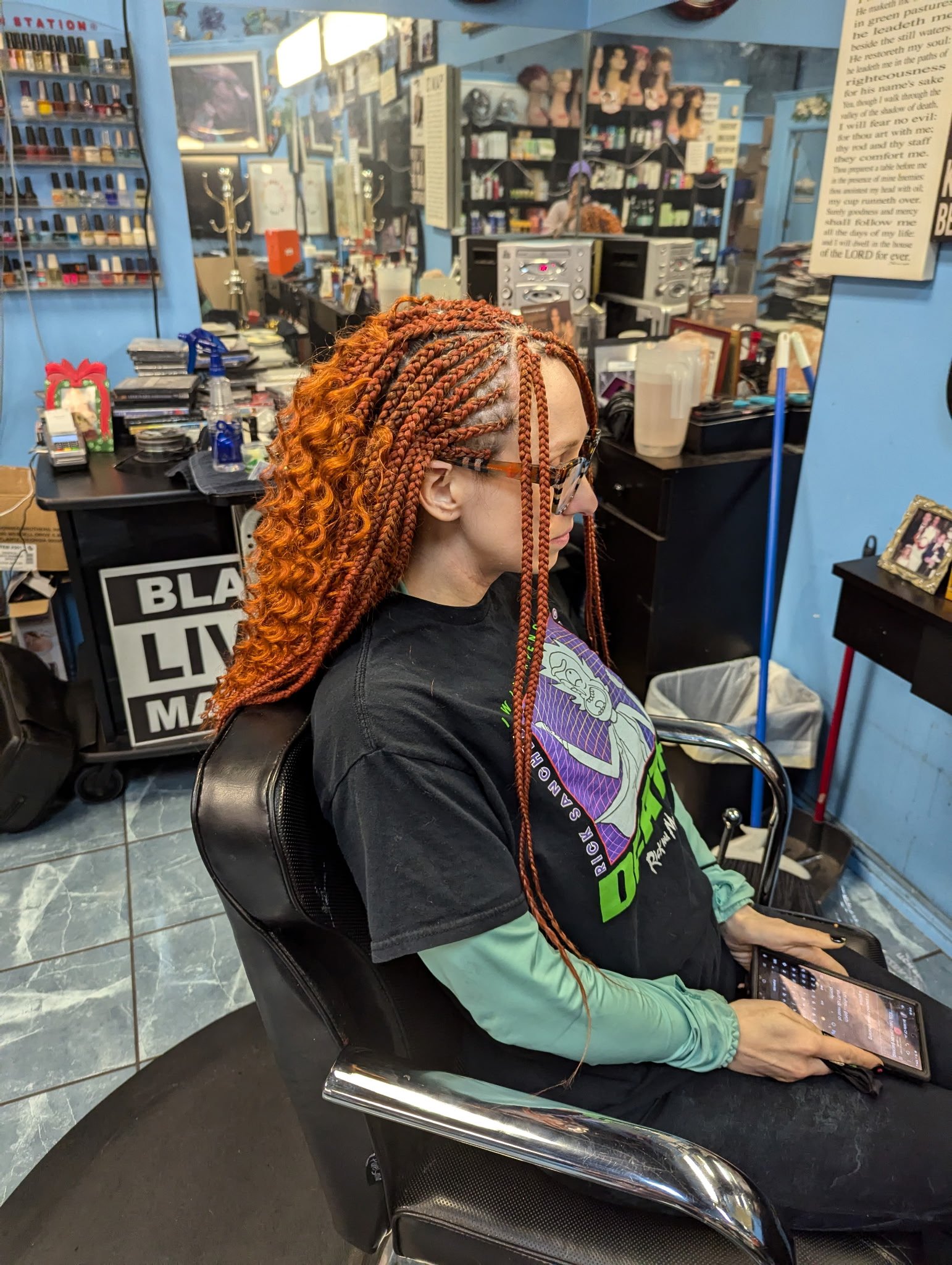 Person with braided orange hair sitting in a salon chair holding a tablet, with salon supplies and decor visible in the background.