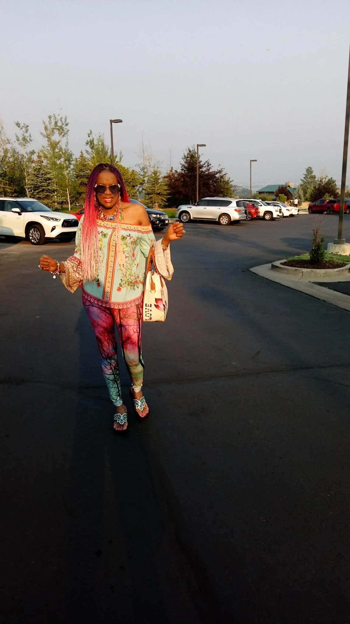 Person with colorful braids and a vibrant outfit standing in a parking lot with cars and trees in the background.