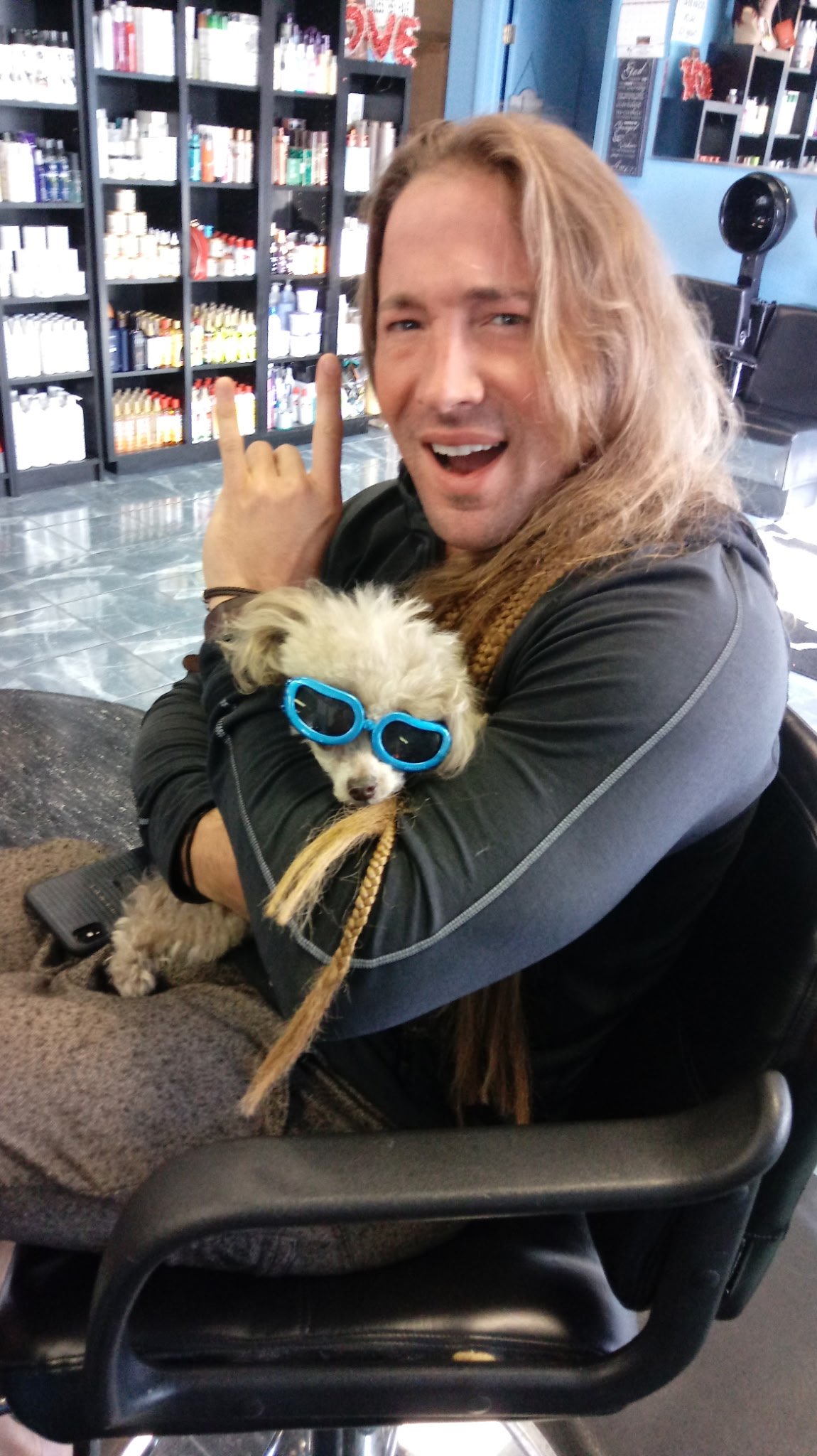 Man with long hair holding a small dog wearing blue sunglasses, sitting in a salon.