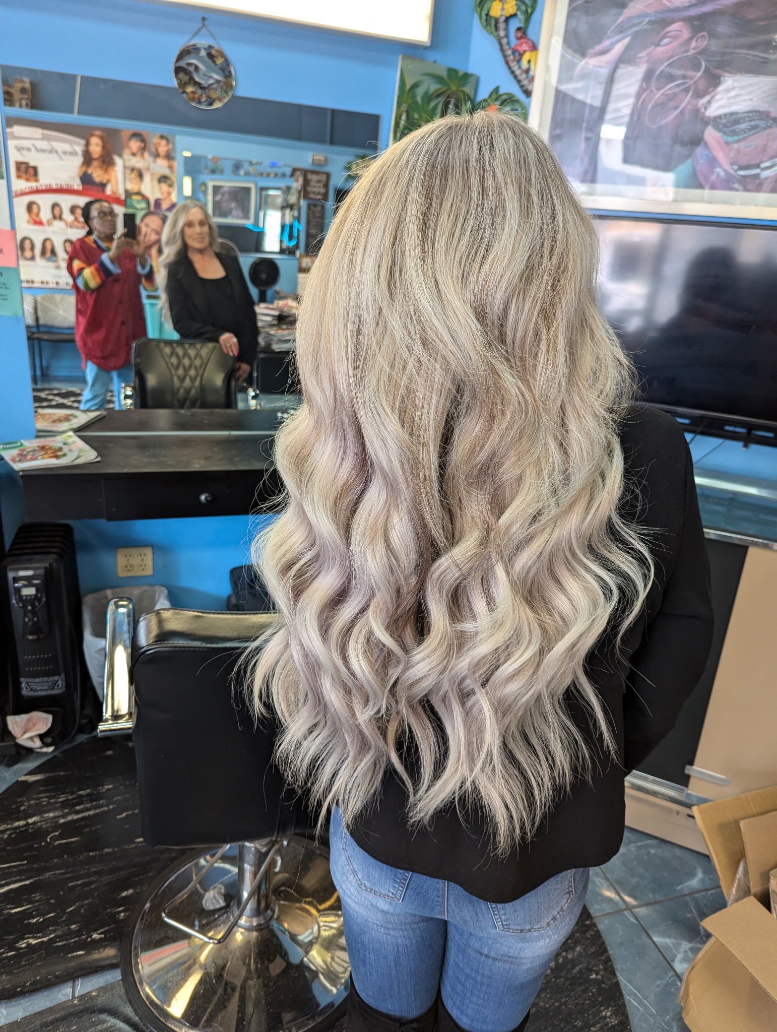 Person with long, wavy blonde hair in salon setting, mirror reflections of people in background.
