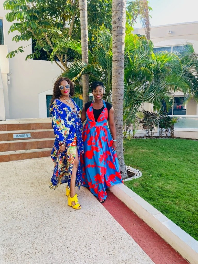 Two women in colorful dresses posing outdoors near palm trees, smiling.