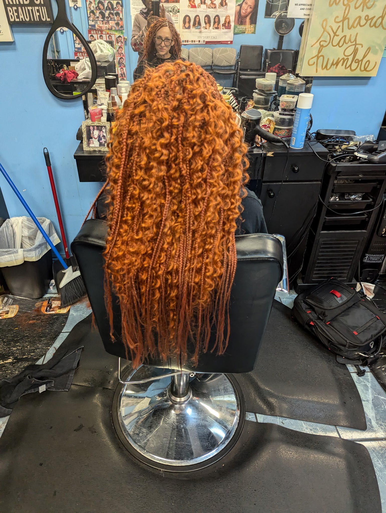 A person with long, curly red hair extensions sitting in a salon chair facing a mirror in a hair salon. The salon is equipped with various hair care products and tools, and there is a decorative sign on the wall.