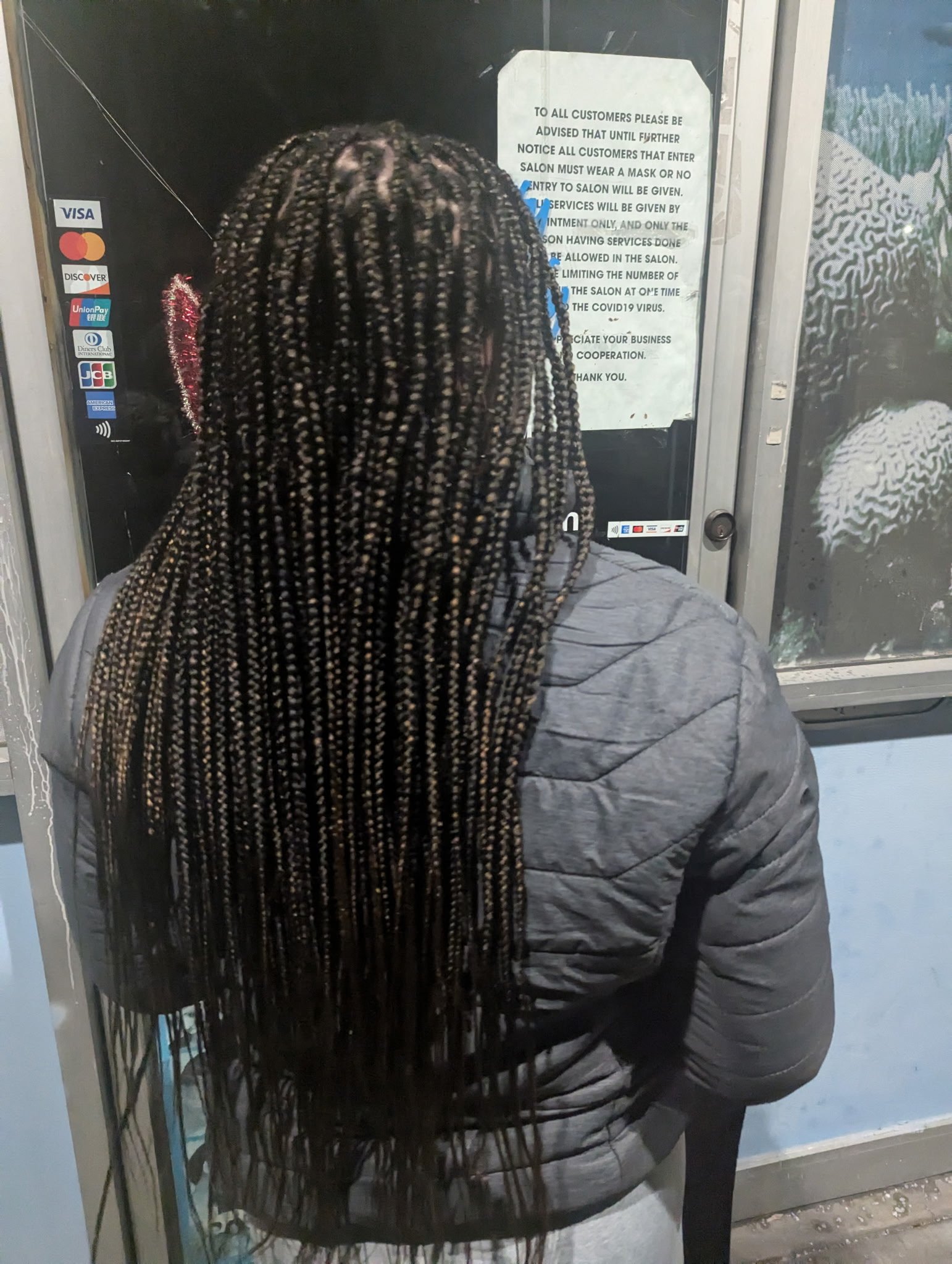 Person with long braided hair wearing a gray puffer jacket standing outside a salon reading a notice on the glass door.