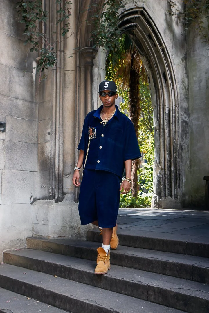 Young man in navy blue outfit, with gold jewelry, sunglasses, cap, and tan boots, walking out of a gothic stone archway surrounded by greenery in daylight.