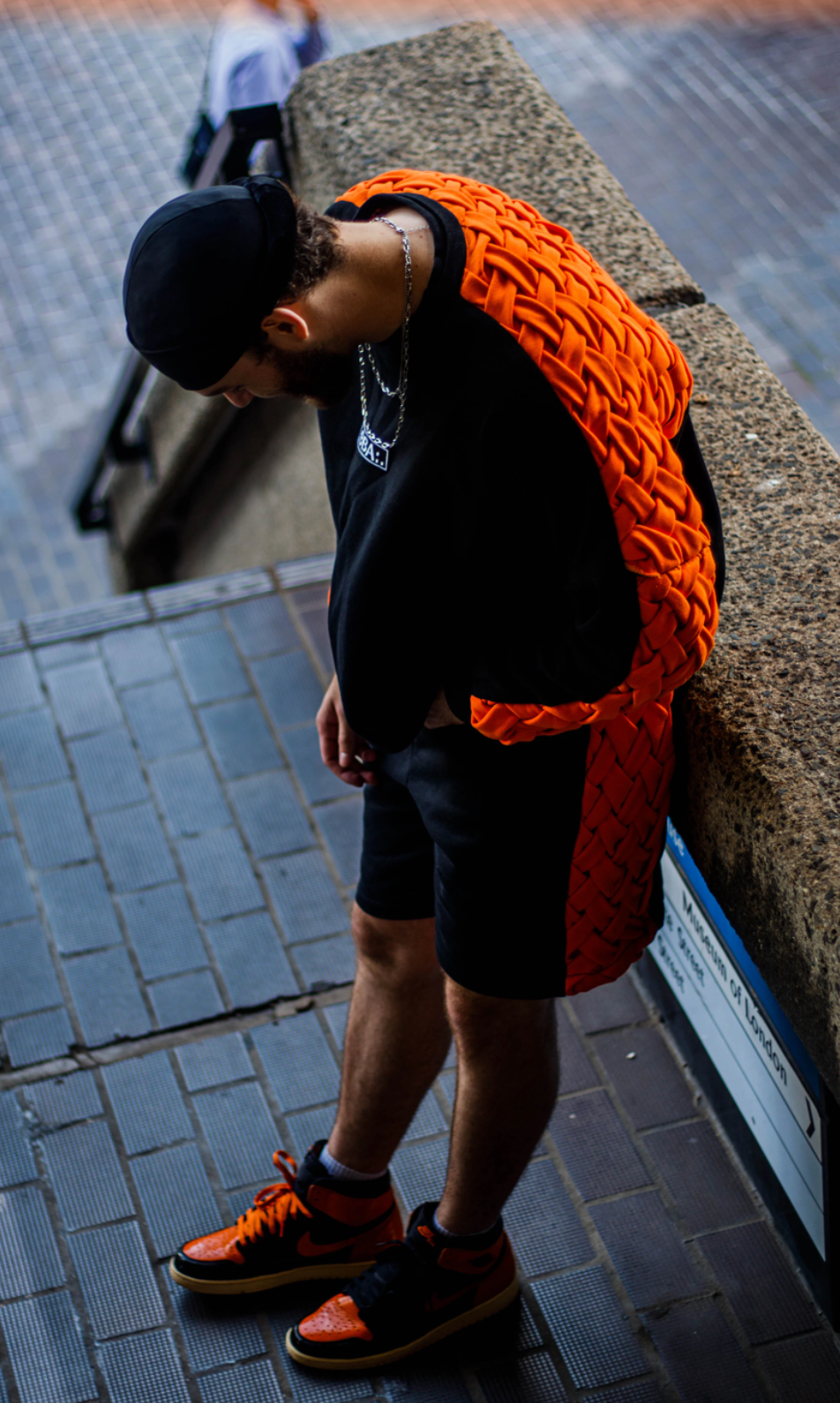 A young man is leaning over a stone ledge on a sidewalk, wearing a black cap, black shirt with a chain necklace, black shorts, and orange and black sneakers. He is carrying an orange woven backpack.