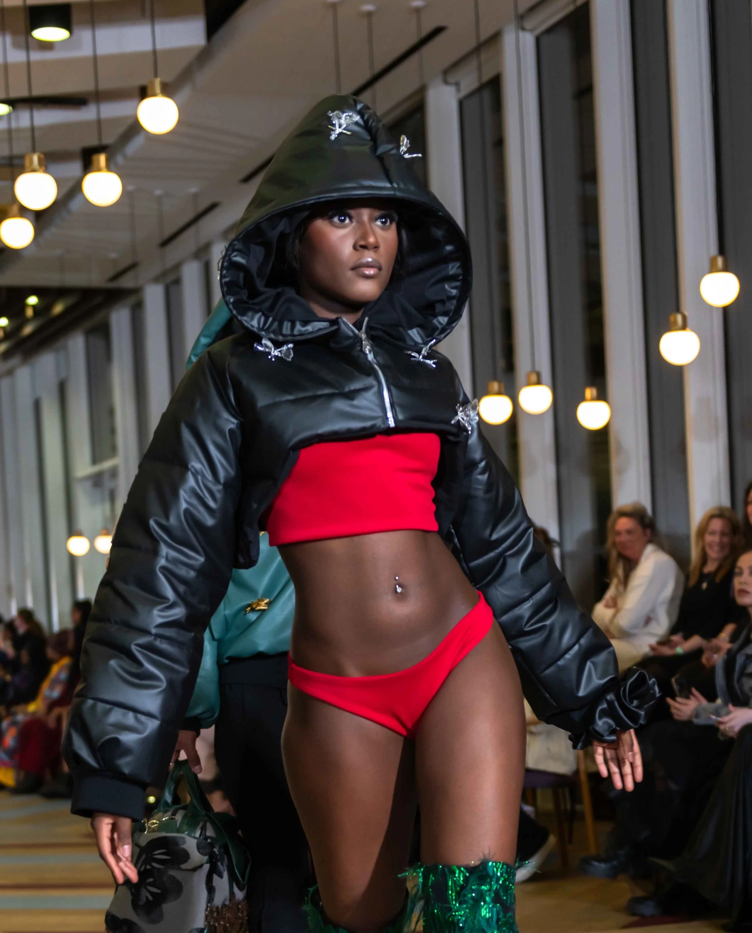 A woman walking runway in red bikini bottoms, black jacket, and green shiny boots at fashion show.