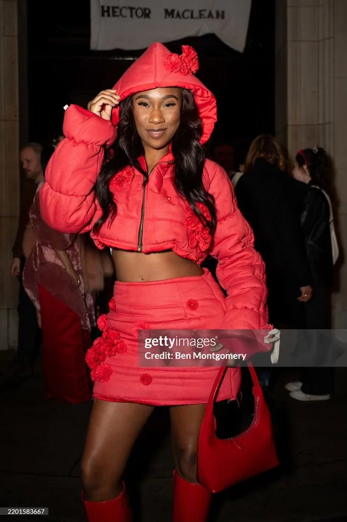 A woman wearing a red cropped puffer jacket with floral accents on the skirt and hood, matching red skirt, and red boots, holding a red bag, standing in a crowded indoor event.
