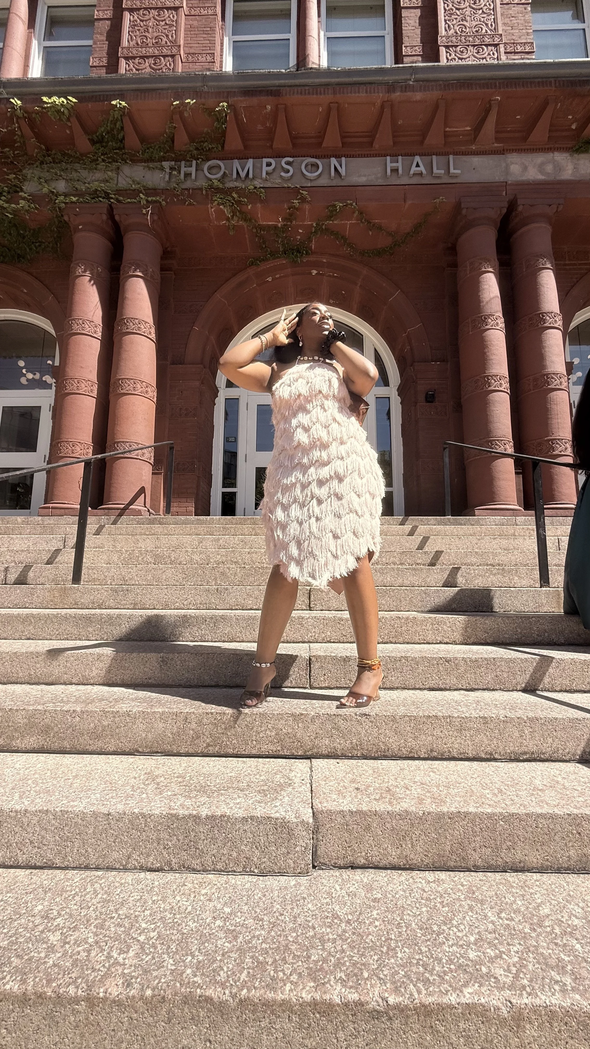 A woman wearing a feathered dress standing on the steps in front of Thompson Hall building with red brick architecture and large columns.