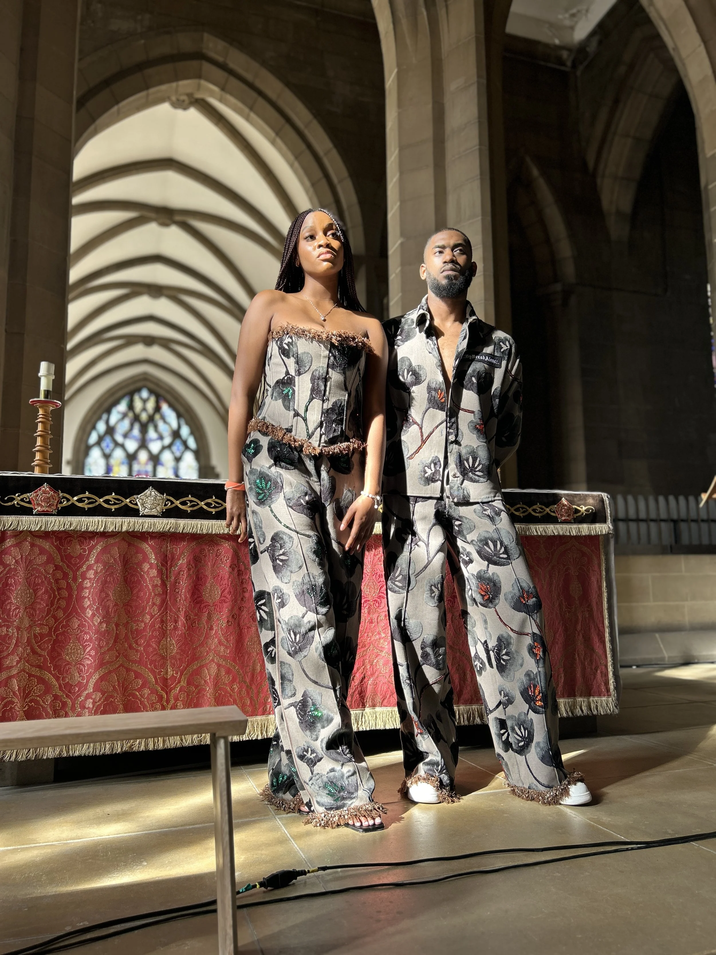Two models standing indoors in front of an ornate table with a red and gold tapestry, in a building with vaulted ceilings and stained glass windows.