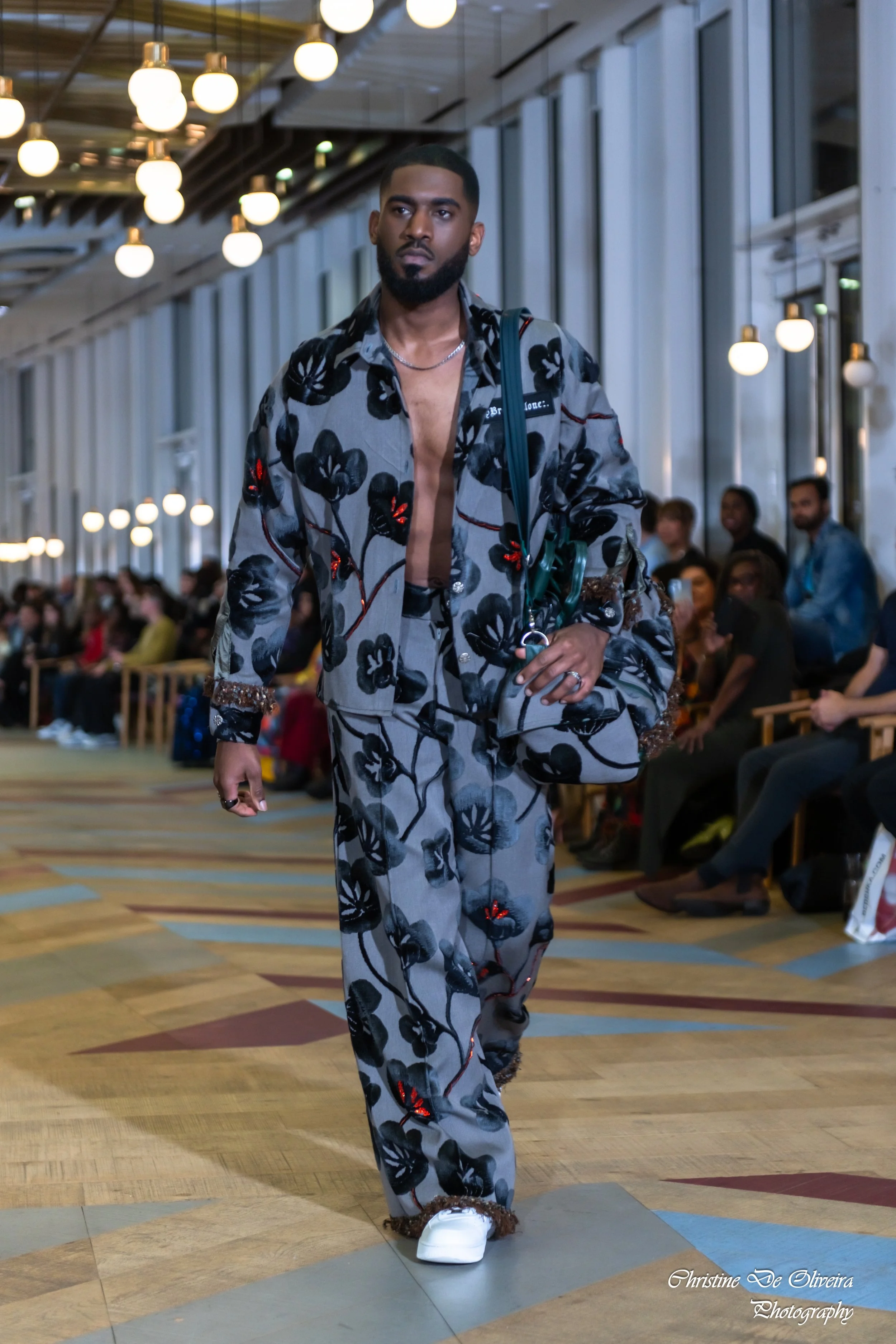 A man with a beard walking down a runway at a fashion show, wearing a matching gray floral suit with black and red accents, and white sneakers with fur trim. There is a seated audience in the background and modern lighting.