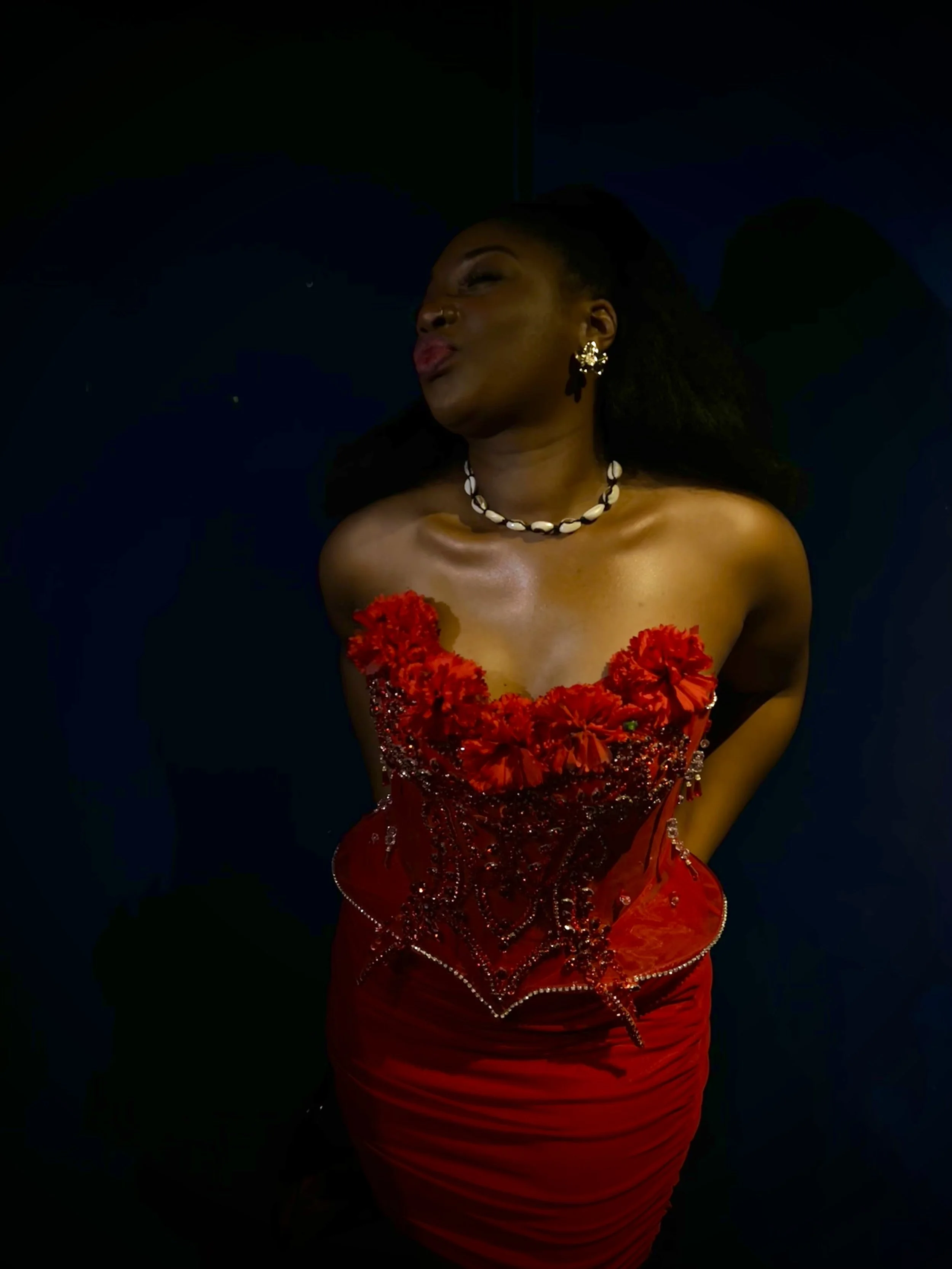 A woman with dark hair in a side profile pose, wearing a red strapless dress with floral embellishments, a shell necklace, and earrings, posing against a dark background.