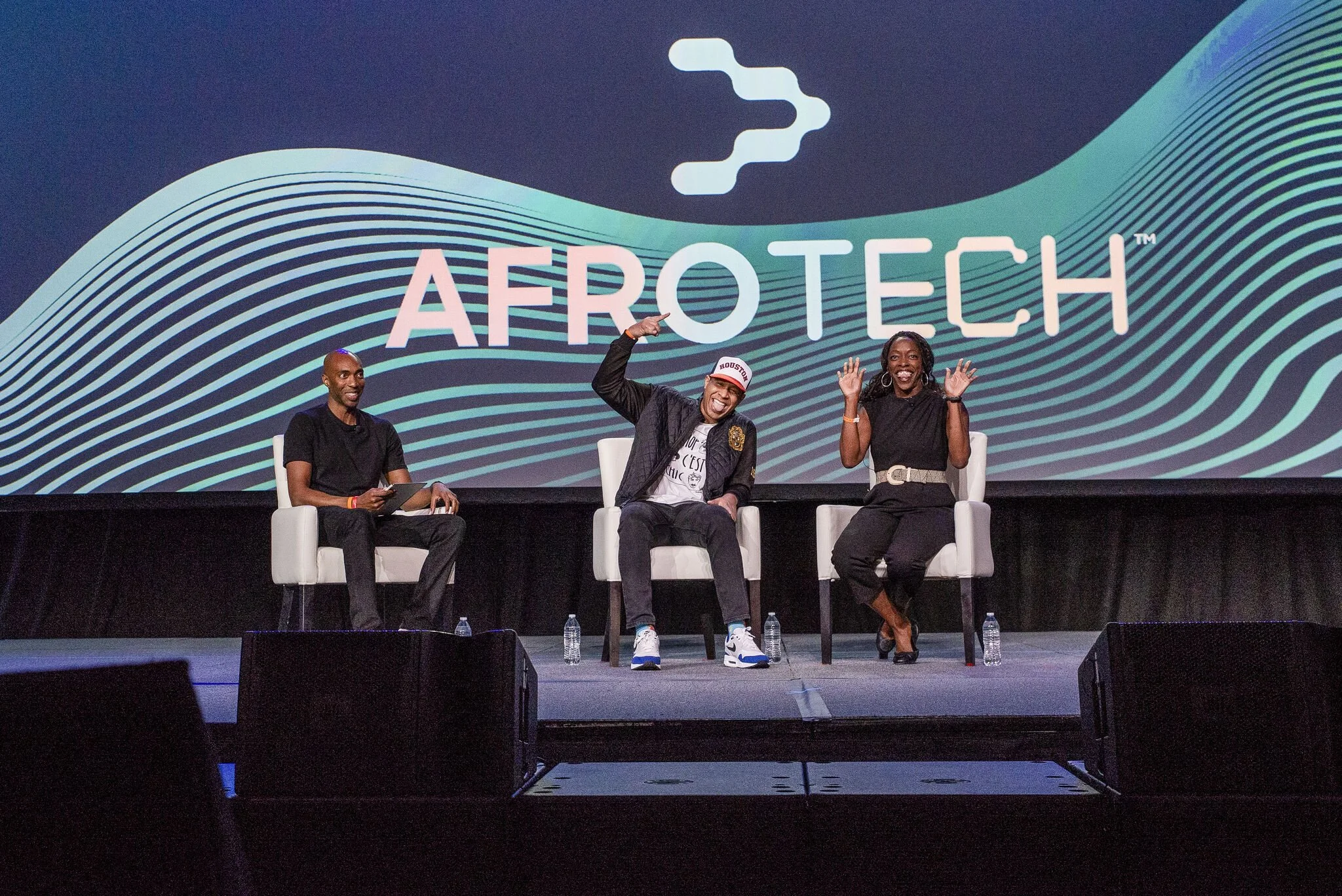 Three people sitting on chairs on stage at an AeroTech event. The backdrop displays the AeroTech logo with a wave pattern. The person in the middle is wearing a white cap and making a peace sign, the person on the right is raising both hands in the air, and the person on the left is holding a phone.