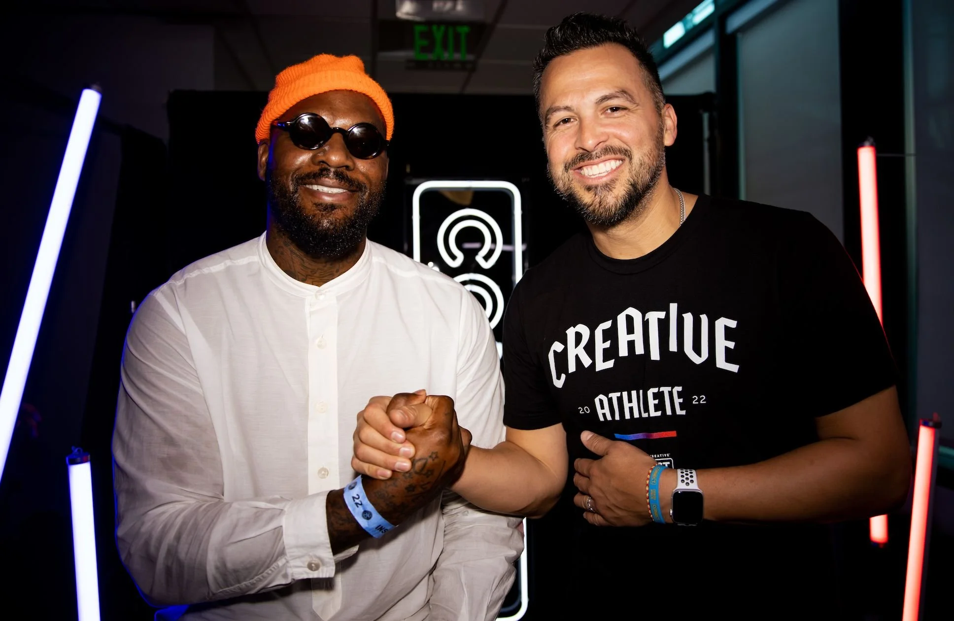 Two men smiling and shaking hands indoors, with neon lights and an exit sign in the background.