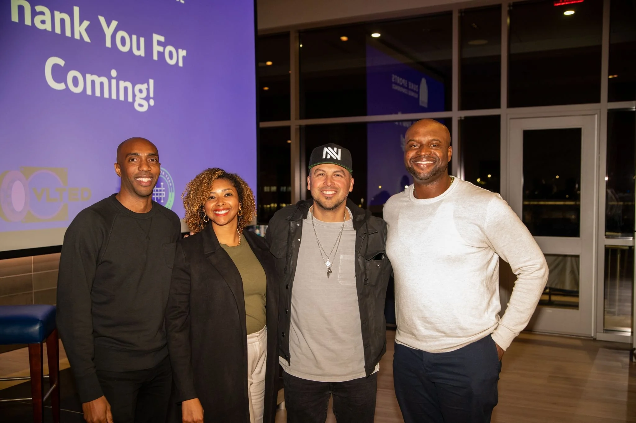 Group of four smiling people standing inside a building, with a large screen behind them displaying a thank you message.