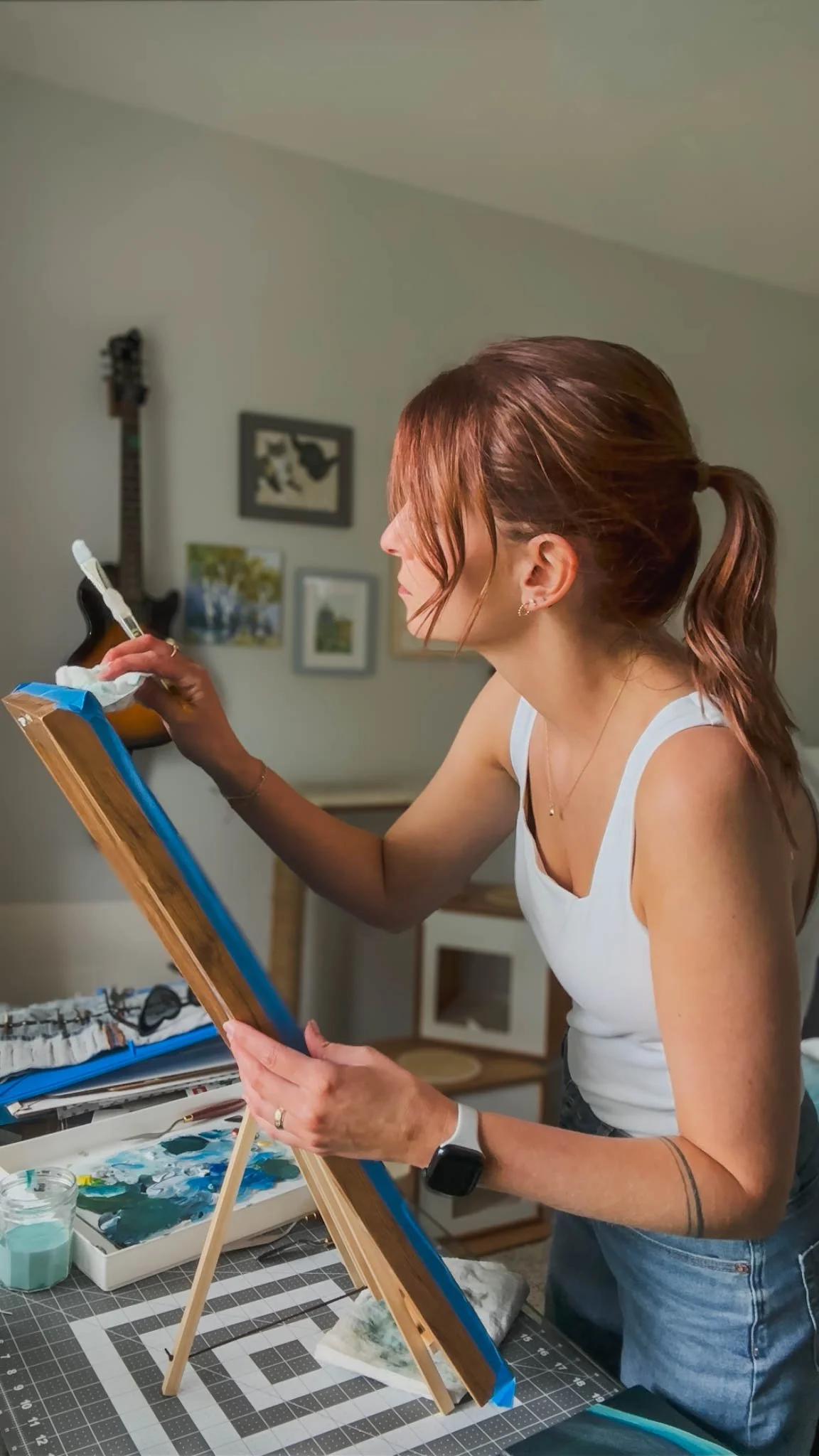 Alissa standing at an easel and mixing paints, with a painting in progress. Art supplies and a palette are on the table, and a guitar hangs on the wall in the background.