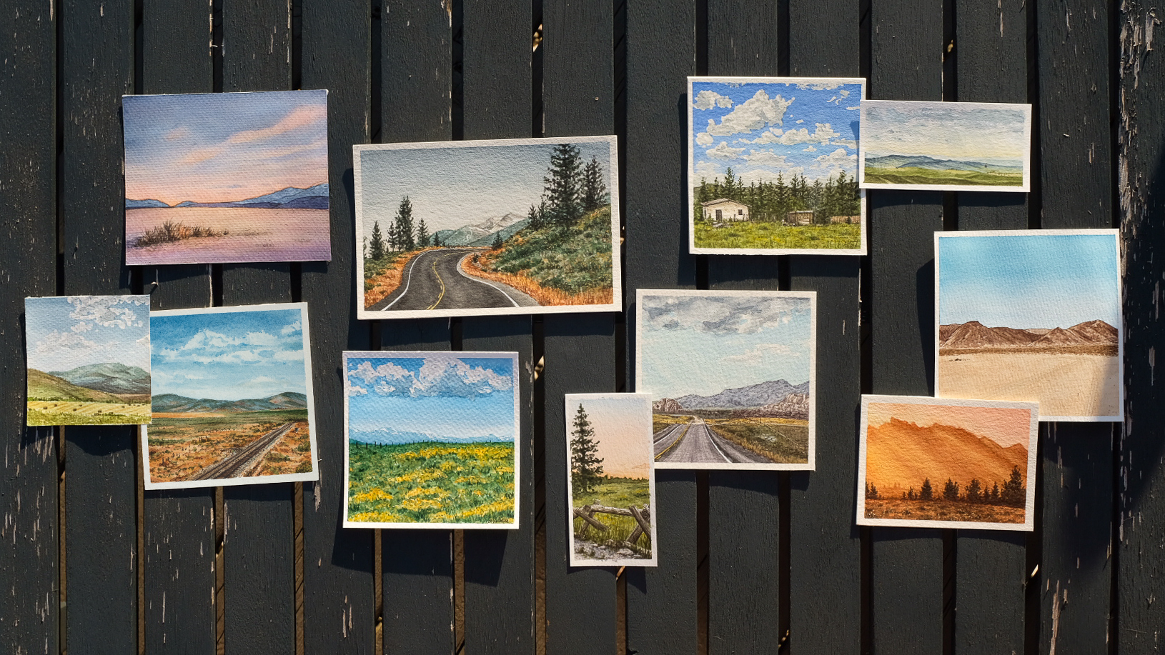 Collection of small landscape watercolor paintings displayed on a wooden fence, depicting mountains, roads, fields, and skies.