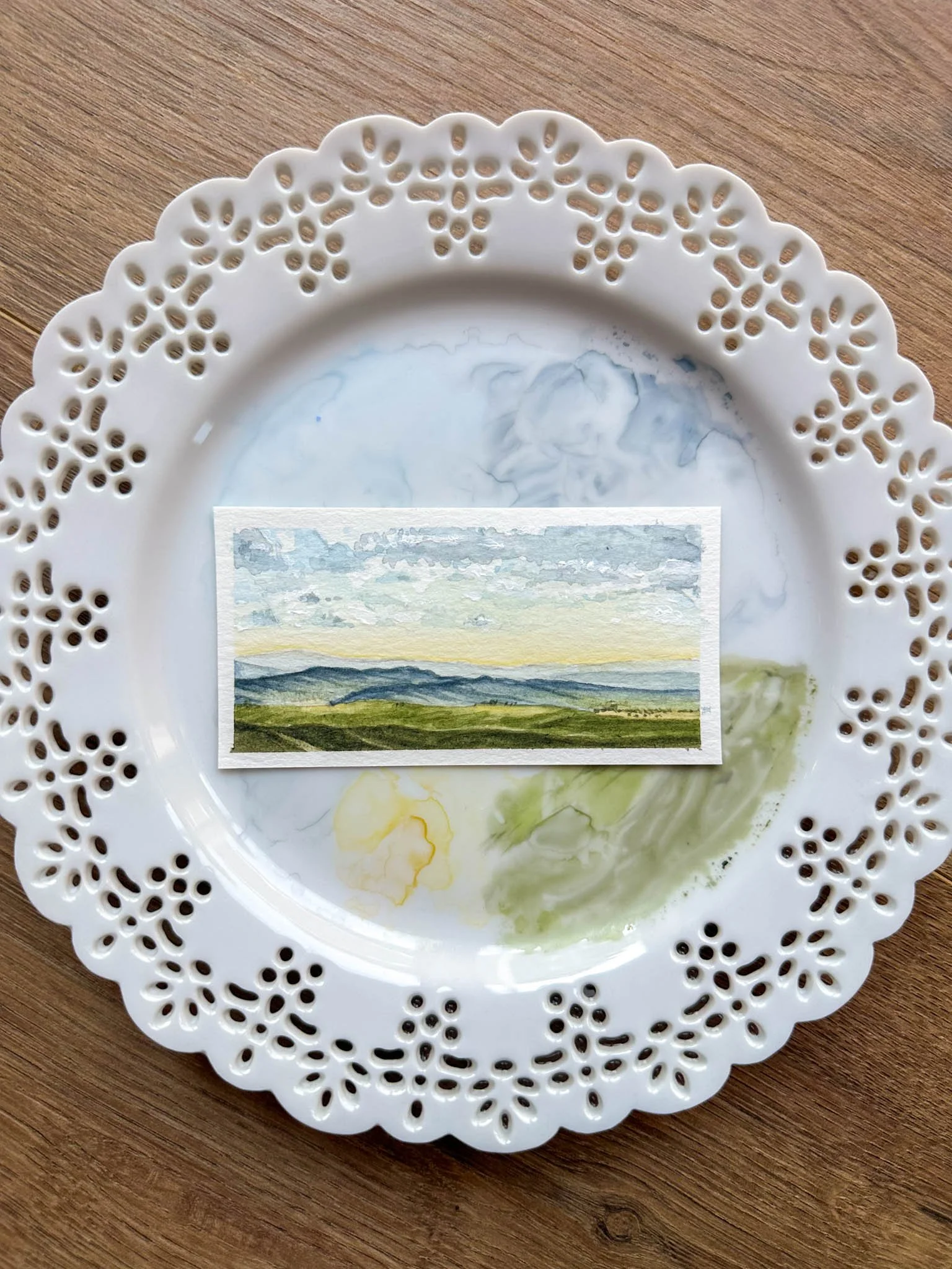 A decorative white plate with intricate cutout patterns around the edges, containing faint watercolor landscape art with hills and a cloudy sky, placed on a wooden surface.