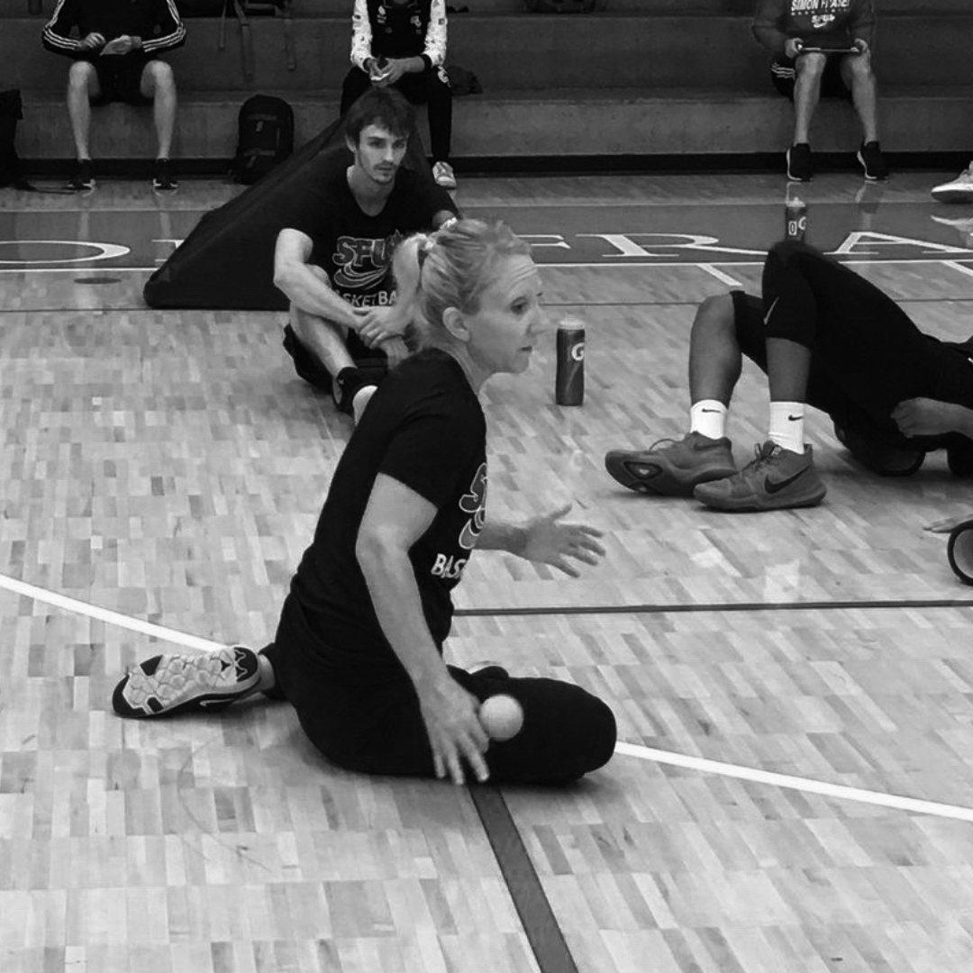Team athletic training session, coach kneeling on gym floor, holding a massage ball, athletes stretching and sitting in background, wooden basketball court surface.
