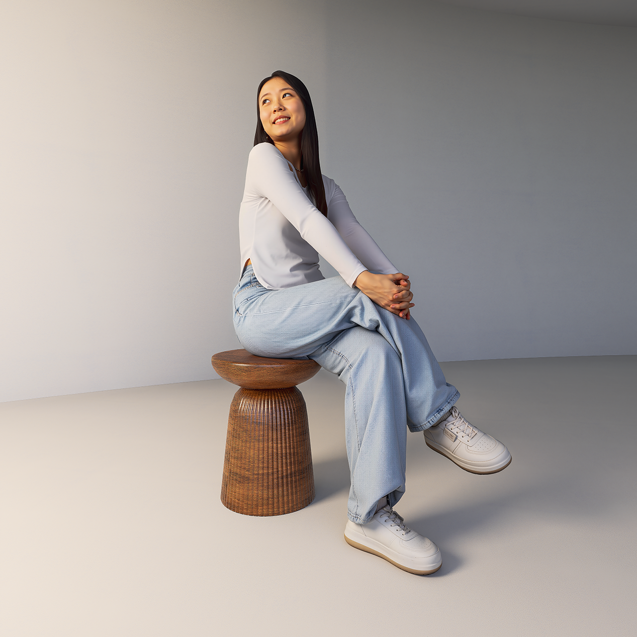 Person sitting on a wooden stool, wearing a white long-sleeve top, light blue jeans, and white sneakers, against a plain background.