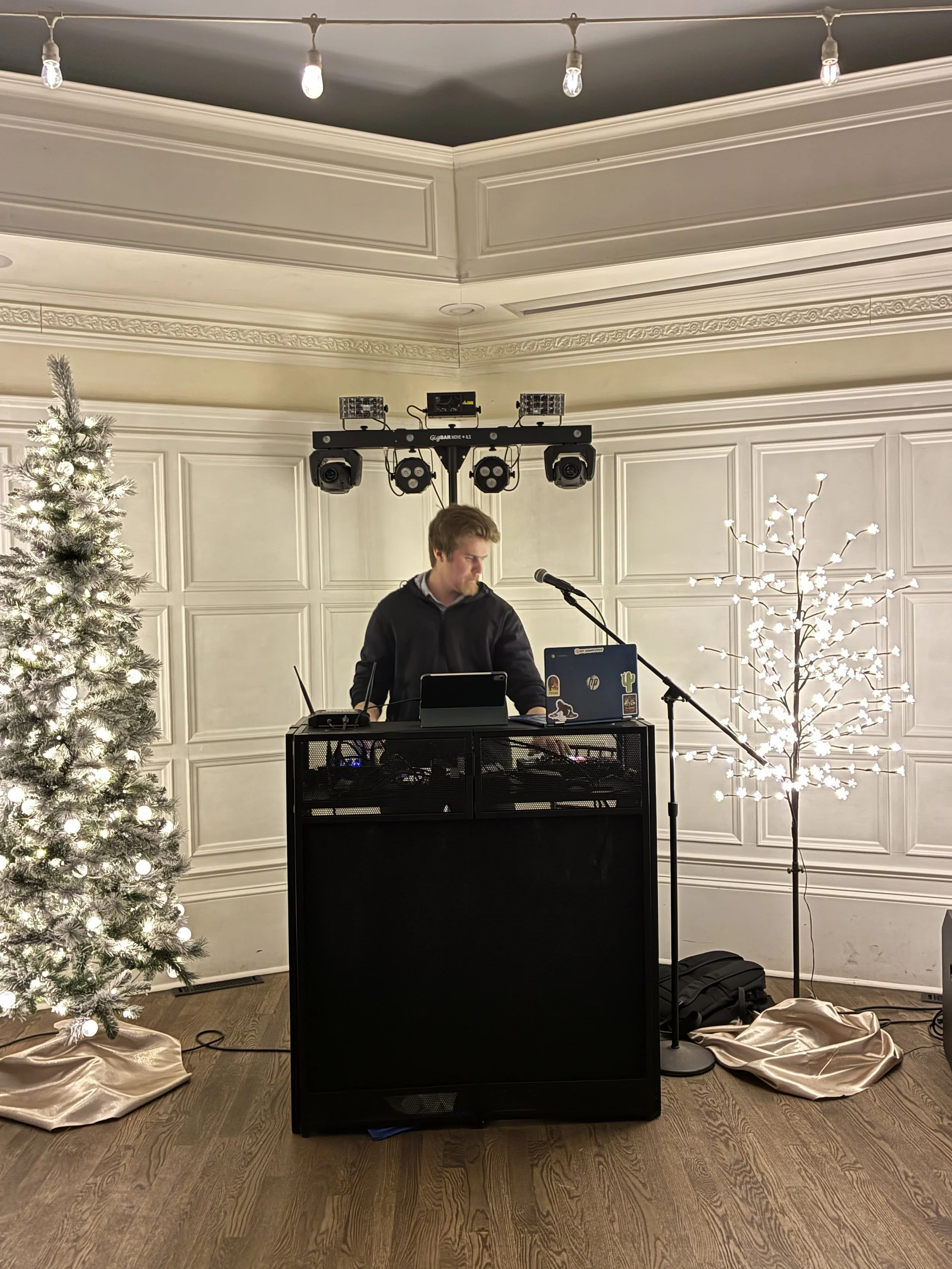Premium DJ Noah, DJ Northh professionally mixing, standing outdoors at night surrounded by trees, working at a DJ booth with DJ equipment, with blue lights and a DJ set up behind him.