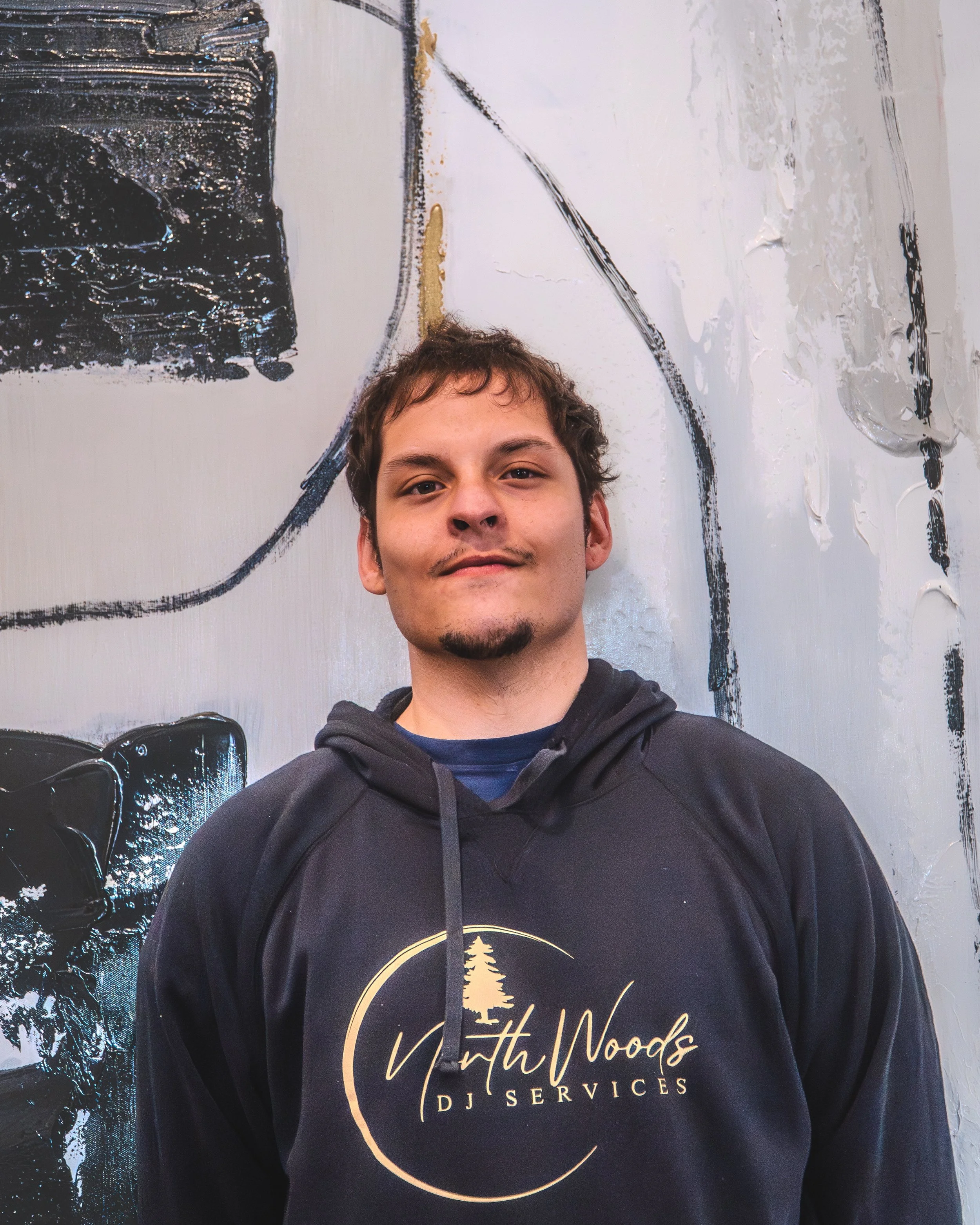 Premium DJ Kenny, better known as DJ Kizz, smiling, wearing a black hoodie, standing against a wooden wall background.