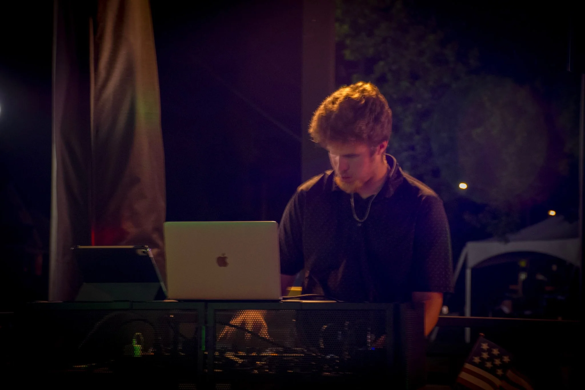 Premium DJ Noah, DJ Northh professionally mixing, standing outdoors at night surrounded by trees, working at a DJ booth with DJ equipment, with blue lights and a DJ set up behind him.