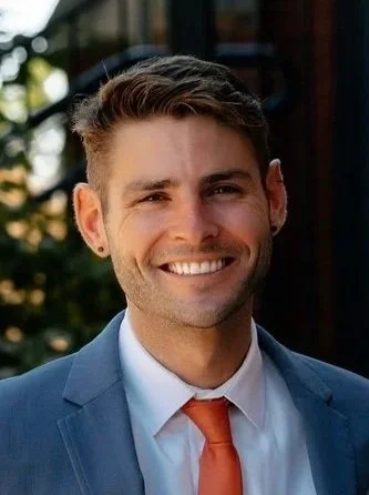 A young man smiling, wearing a blue suit, white shirt, and orange tie, outdoors with blurred greenery in the background.