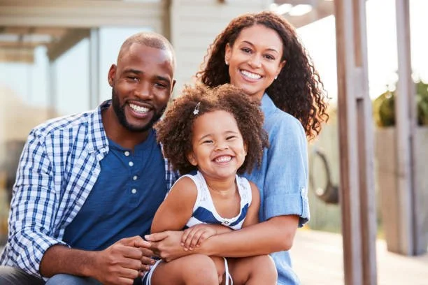 Family of three smiling outdoors, with a father, mother, and young daughter sitting together.