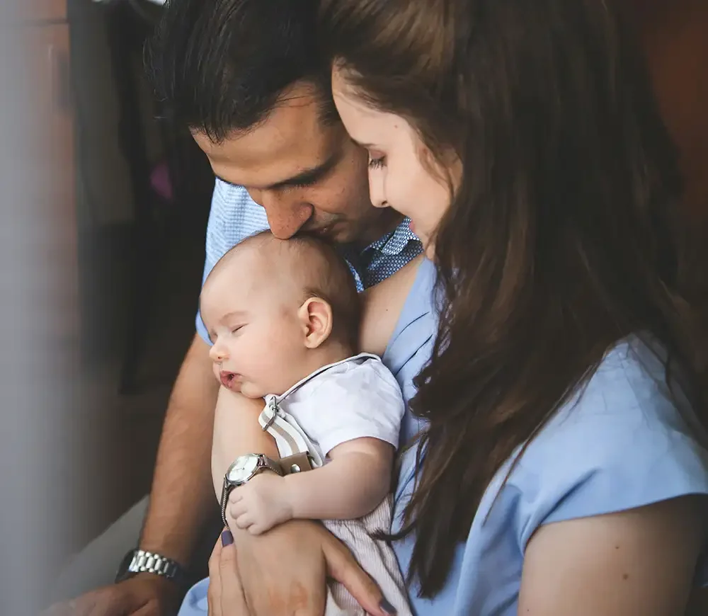 A man and woman hold a sleeping baby close between them, touching foreheads in a tender moment.