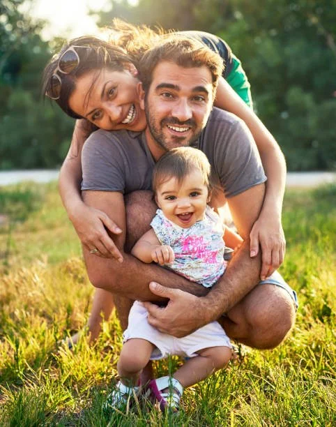 A happy family of three—mother, father, and baby girl—enjoys a sunny day outdoors in a grassy field, with trees in the background. The mother is hugging the father from behind, and the father is holding the baby girl, all smiling at the camera.