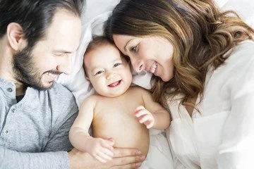 A happy family lying together in bed, smiling and looking at their young child.