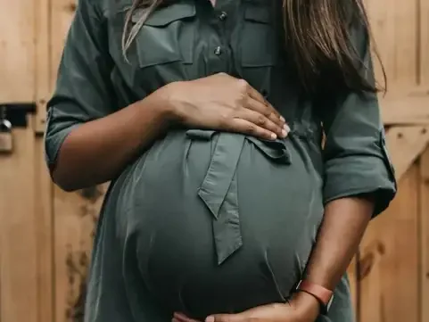 Pregnant woman in green shirt holding her belly with one hand and supporting her belly with the other, standing in front of a wooden background.
