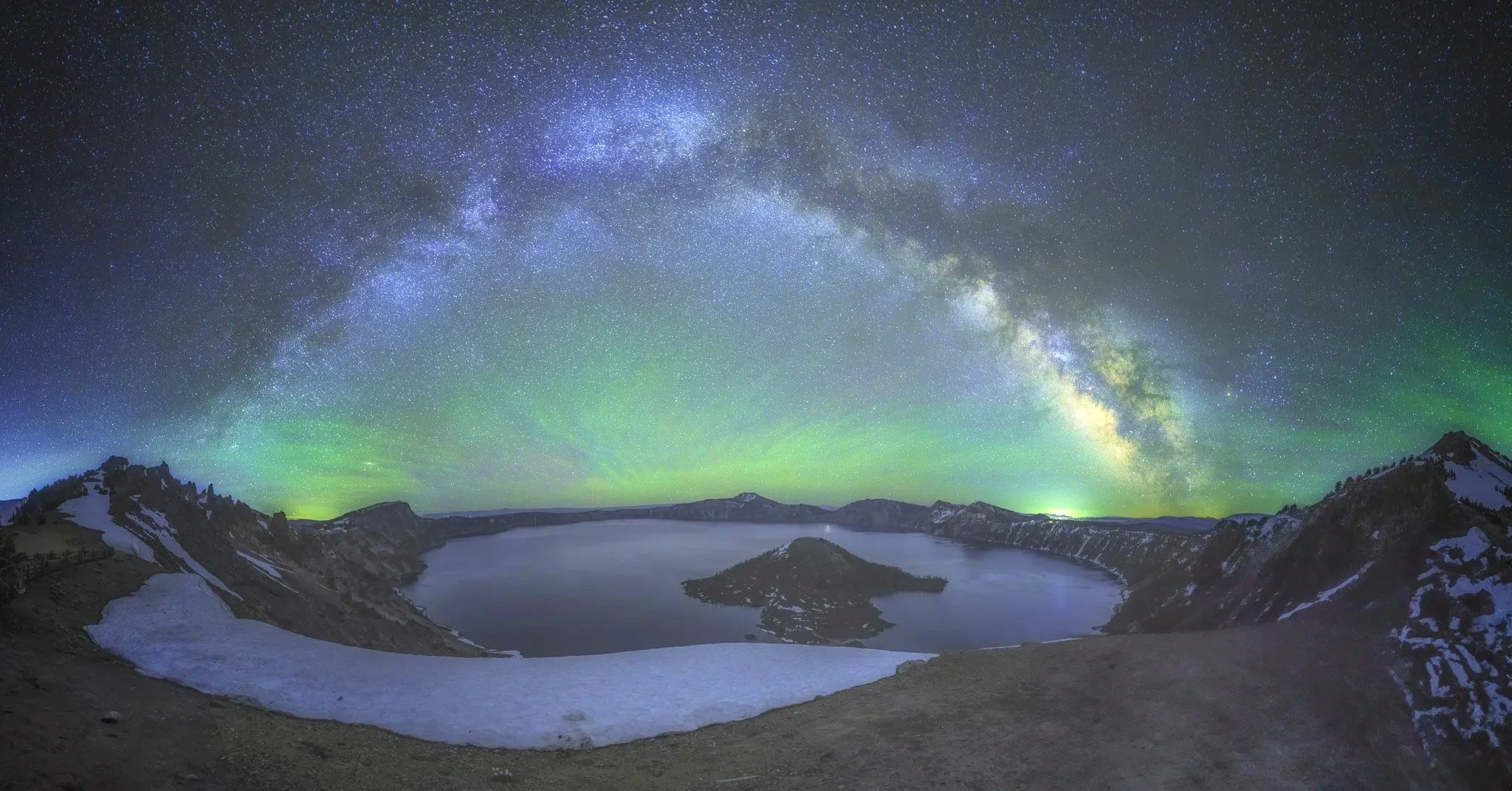 Milky Way arching over Crater Lake with green aurora glowing along the horizon in Oregon.