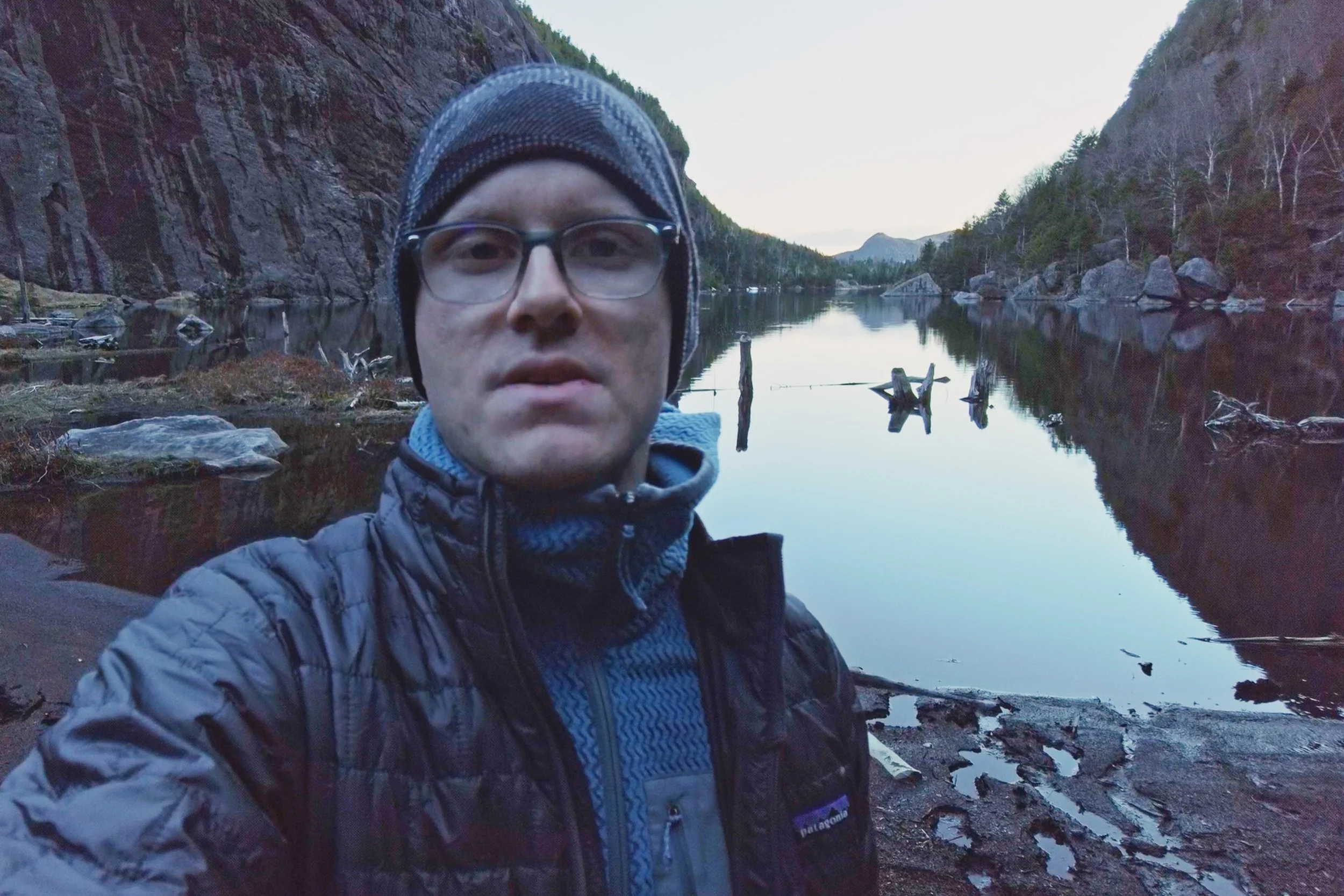 Hiker standing near the shoreline of Avalanche Lake in the Adirondack High Peaks, surrounded by steep cliffs and calm reflective water at dusk.