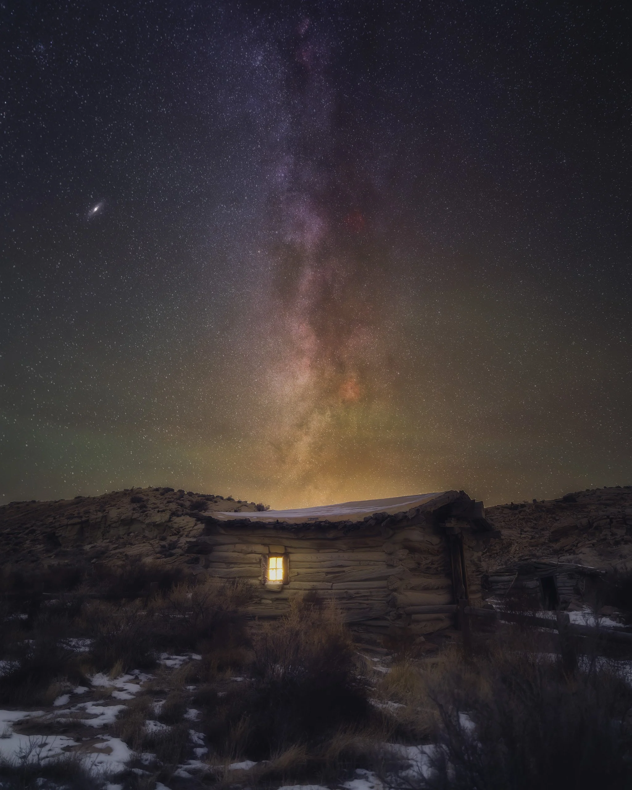 Milky Way rising above Wolfe Ranch Cabin in Arches National Park, Utah, with snow patches on the ground and a glowing window in the rustic log cabin, photographed by Matt Costanza.