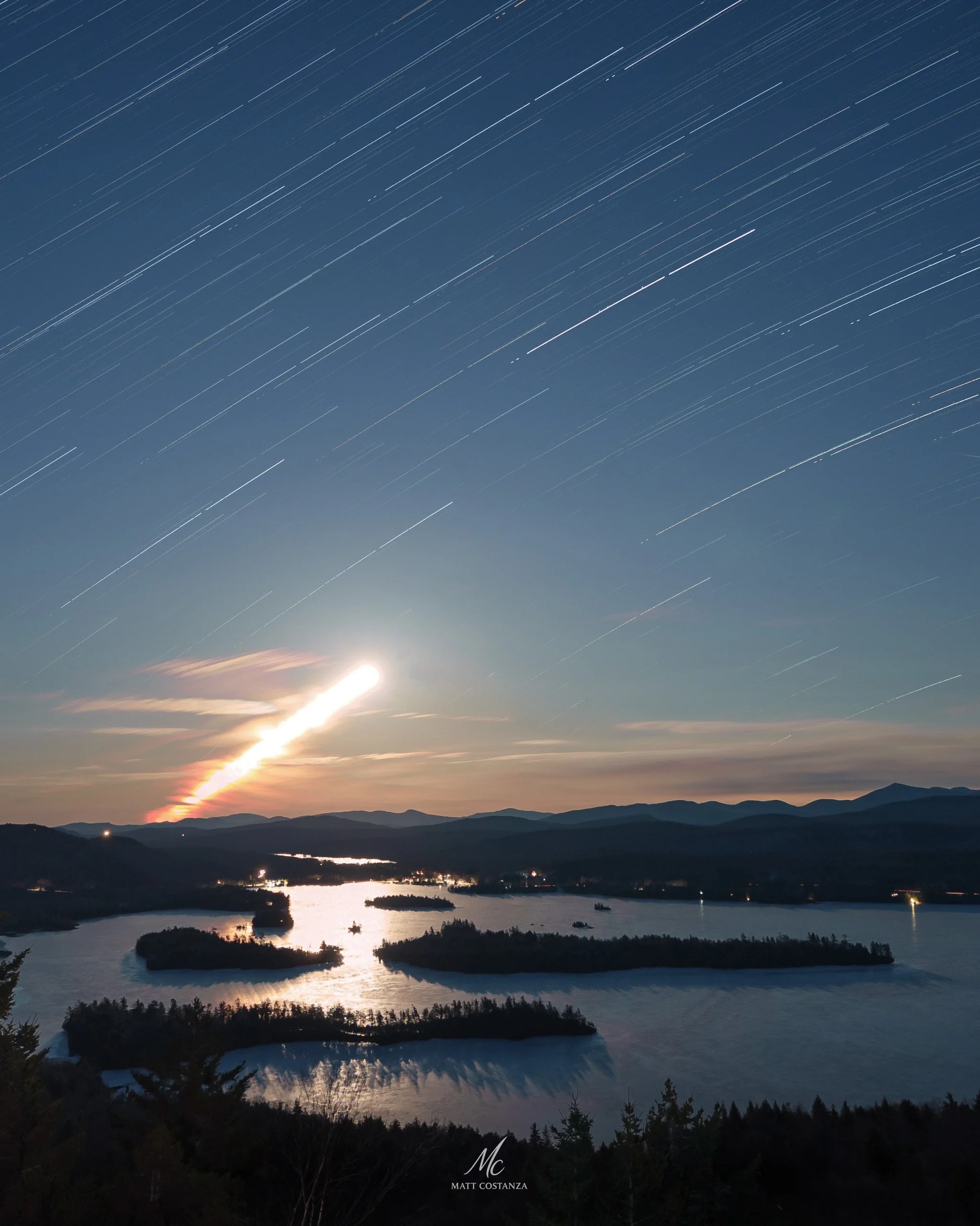 Long-exposure photograph showing a moon trail and star trails over frozen Adirondack lakes and mountain silhouettes at night.