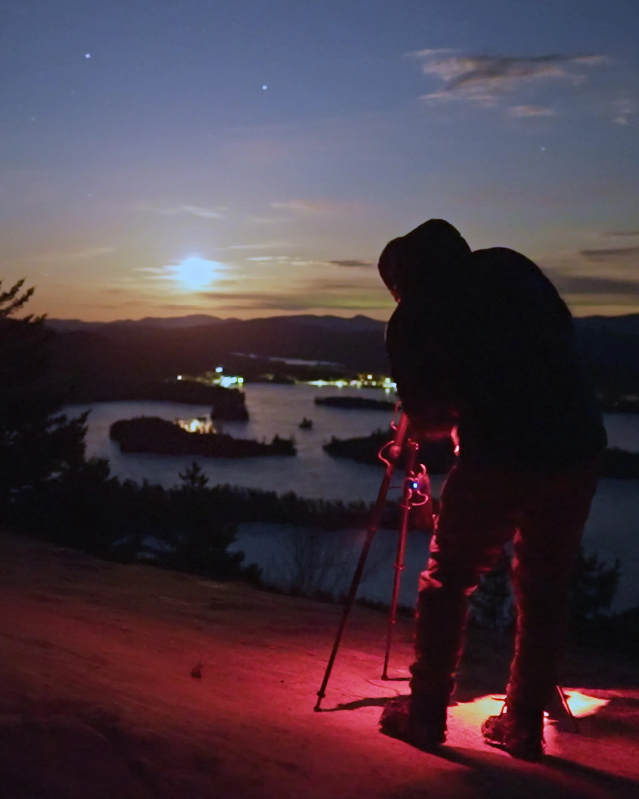 Photographer setting up a tripod to capture a moonrise over Adirondack lakes, illuminated by moonlight and distant shoreline lights.