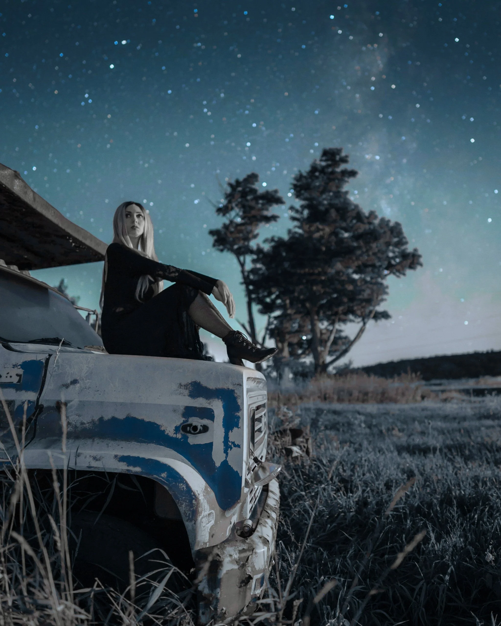Woman sitting on top of a weathered blue truck under the Milky Way in rural Western New York, with trees, grass, and a starry sky in the background, photographed by Matt Costanza.