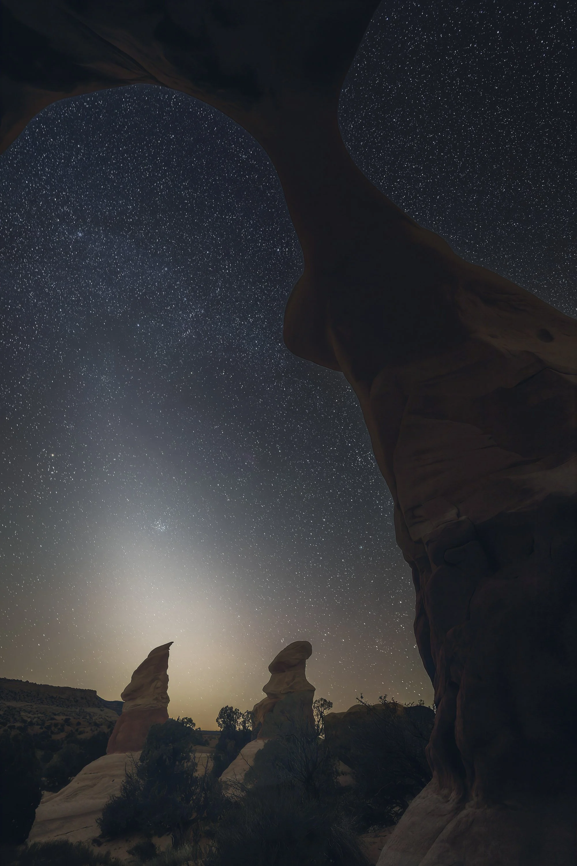 Zodiacal light rising between distant hoodoos beneath Metate Arch at Devils Garden in Grand Staircase-Escalante National Monument, Utah, under a star-filled night sky.