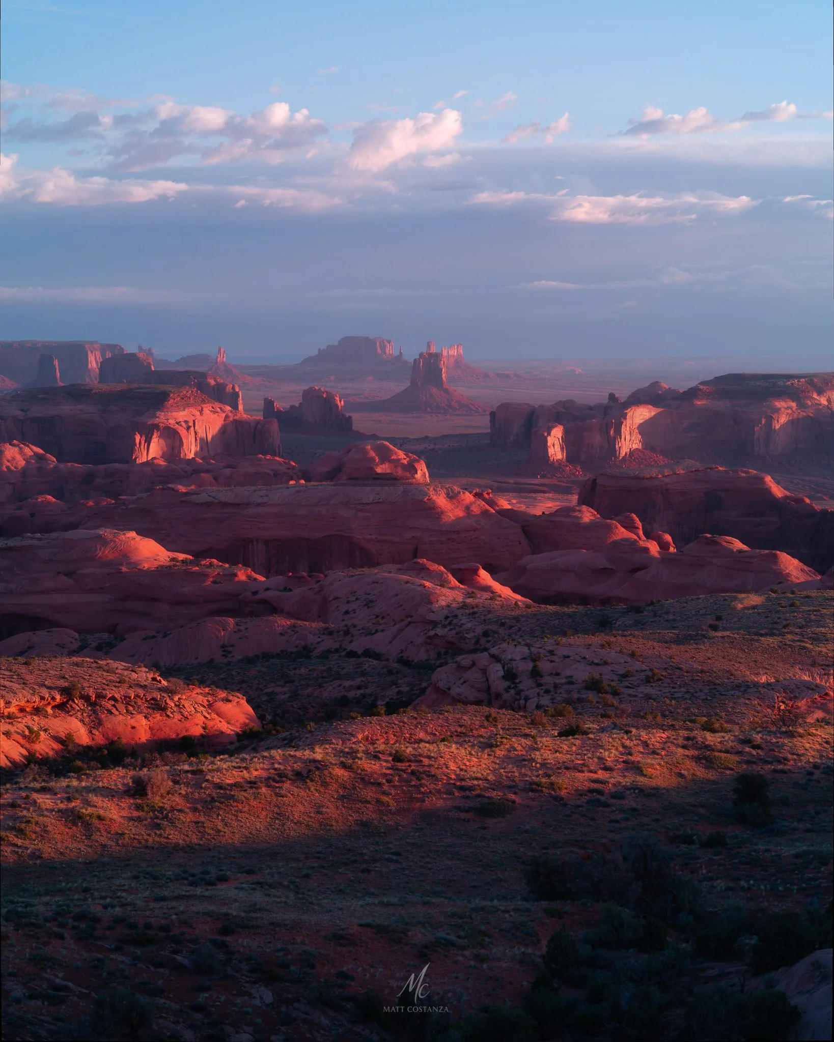 A panoramic sunrise view from Hunts Mesa overlooking Monument Valley’s sandstone buttes and mesas glowing in warm desert light under scattered clouds.