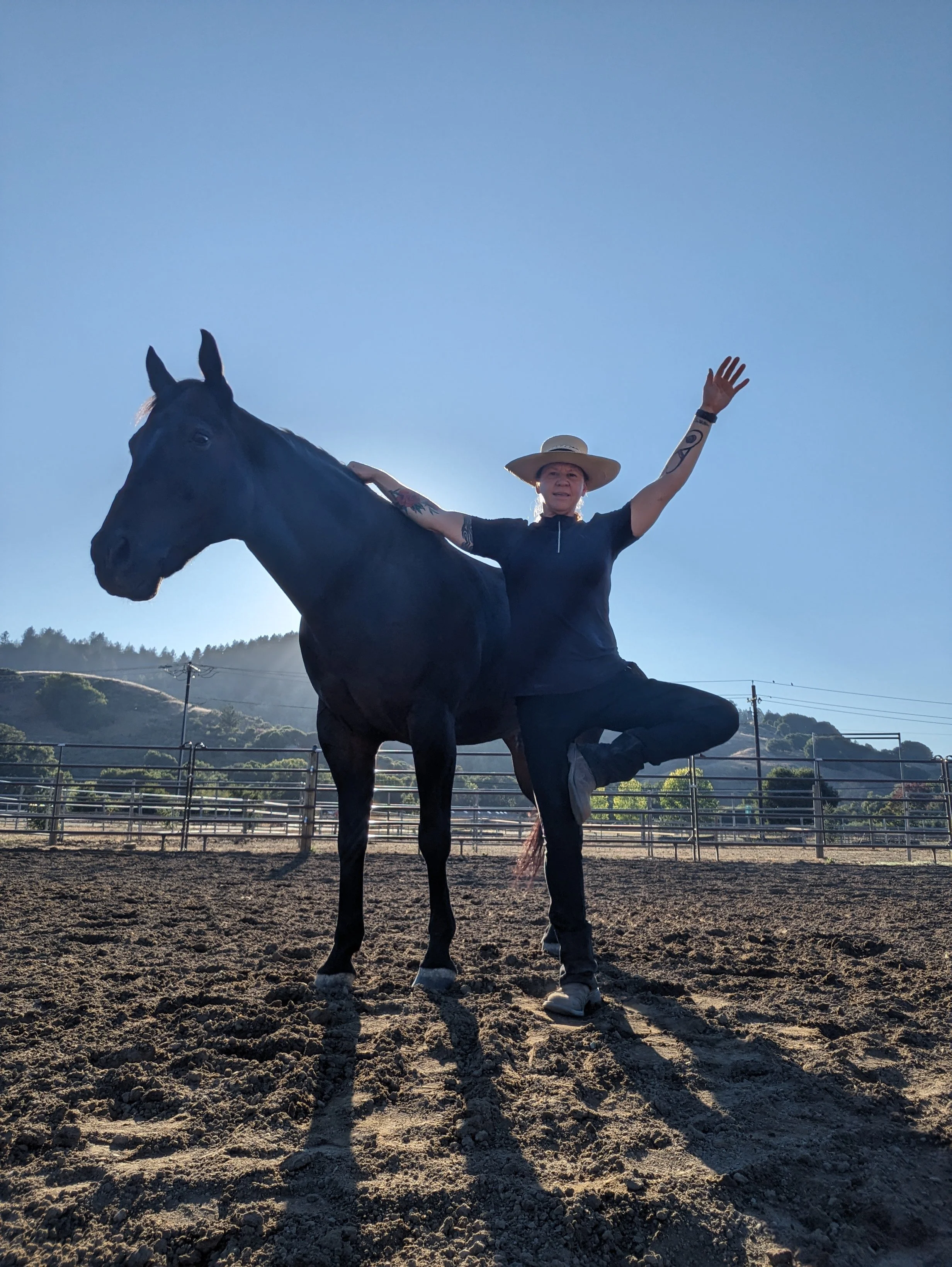 Person standing on one leg with arms raised, next to a black horse in an outdoor riding arena, with hills and a clear blue sky in the background.