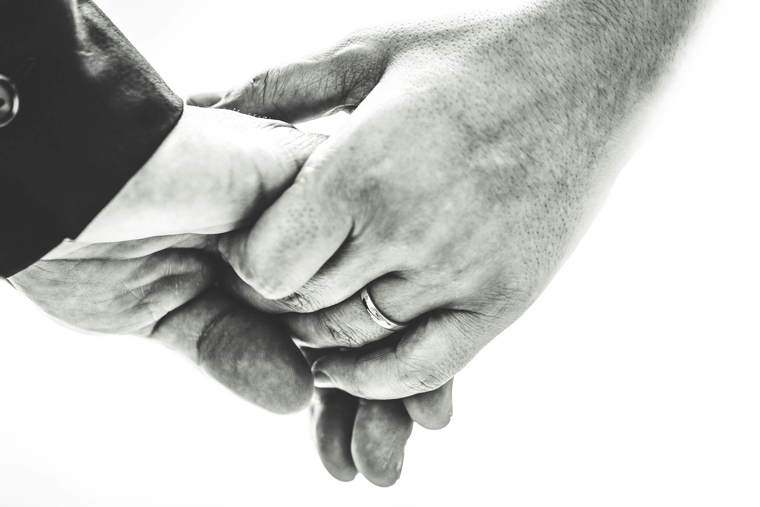 Close-up of two hands holding each other, one with a wedding ring, black and white photograph.