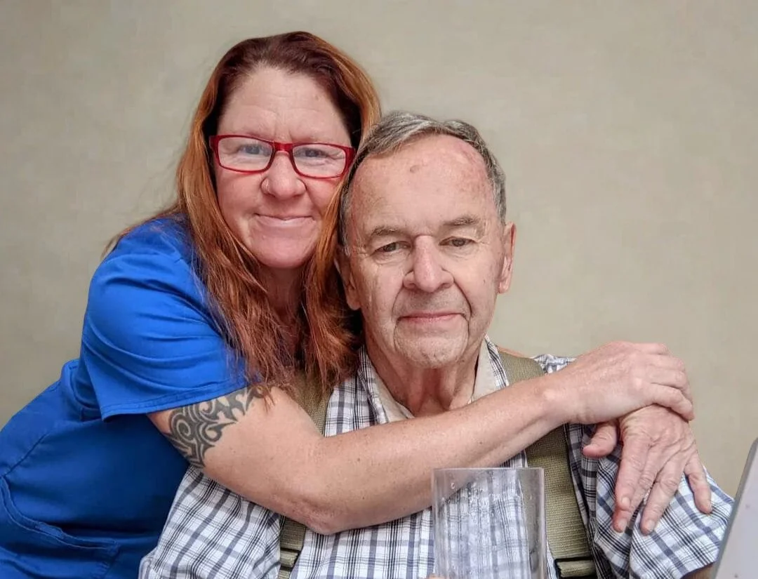 A woman (Melanie Sheaves) with red hair, glasses, and a blue shirt hugging her father in a plaid shirt and suspenders, sitting at a table with a glass and a laptop.