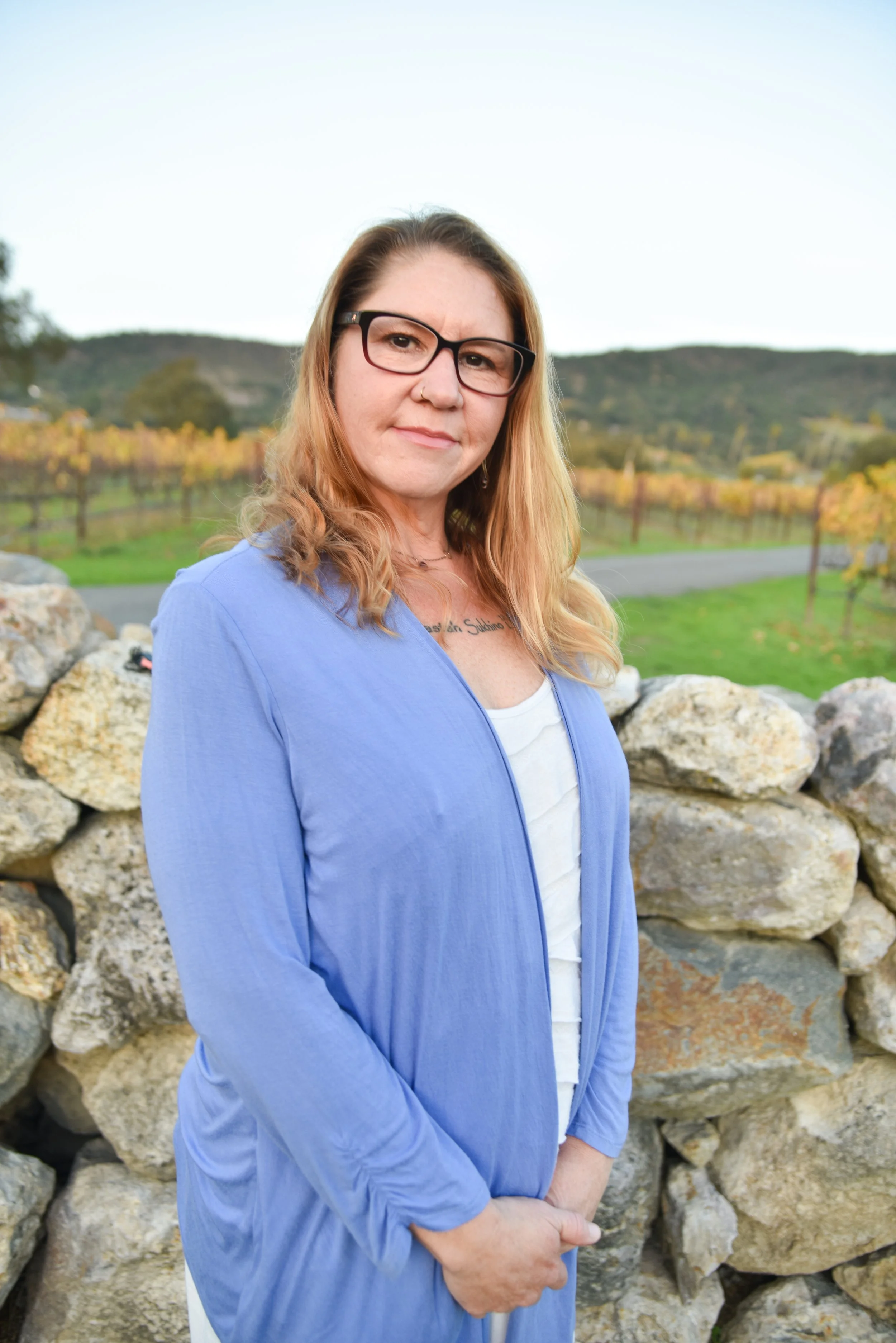 A woman with glasses and wavy red hair standing in front of a stone wall, with a vineyard and hills in the background.