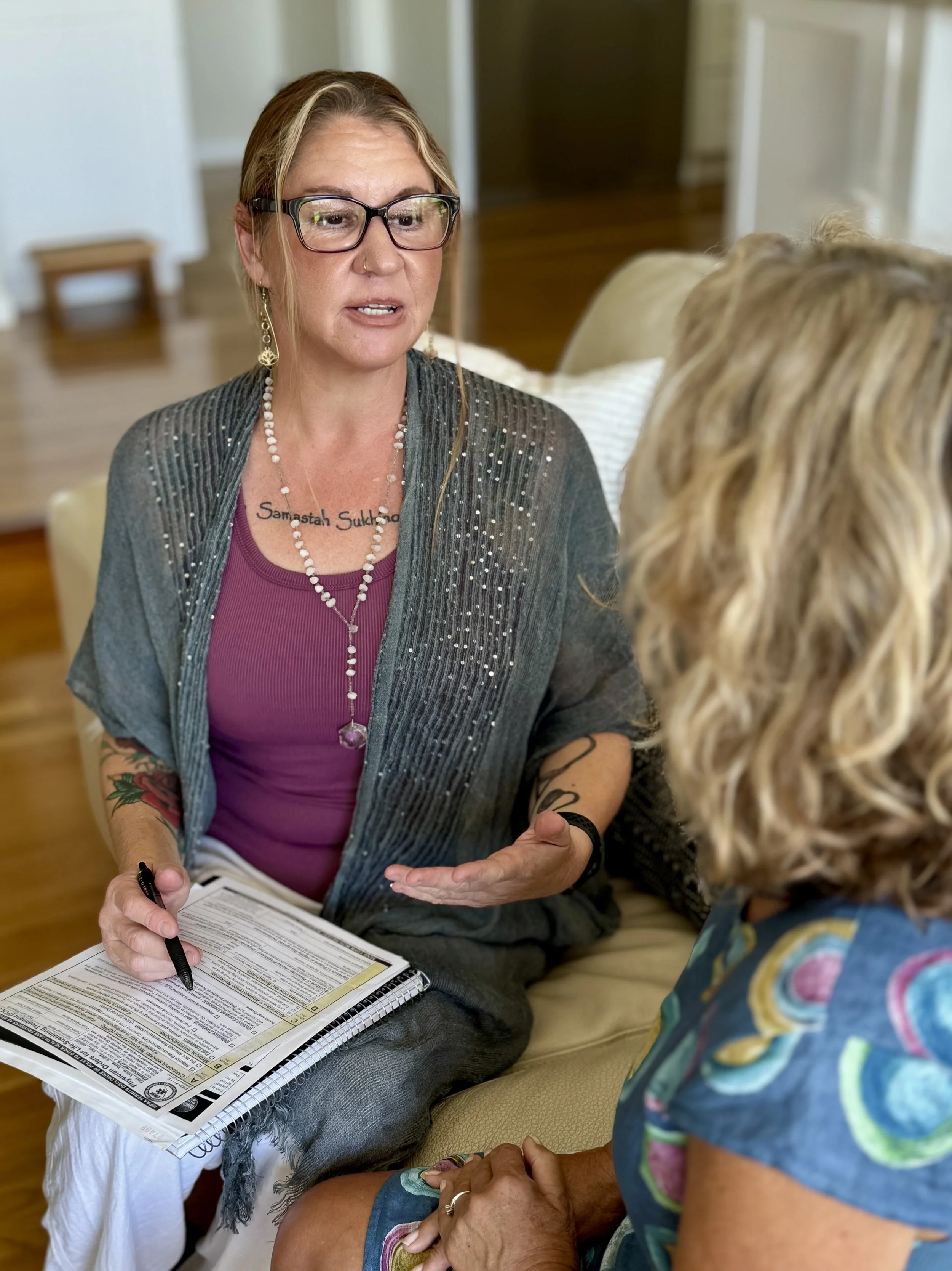 A woman with glasses and tattoos, wearing a purple shirt and a gray cardigan with rhinestones, sitting on a beige sofa, talking to another woman with blonde curly hair, wearing a colorful outfit. The woman with glasses is holding a pen and a clipboard with documents.