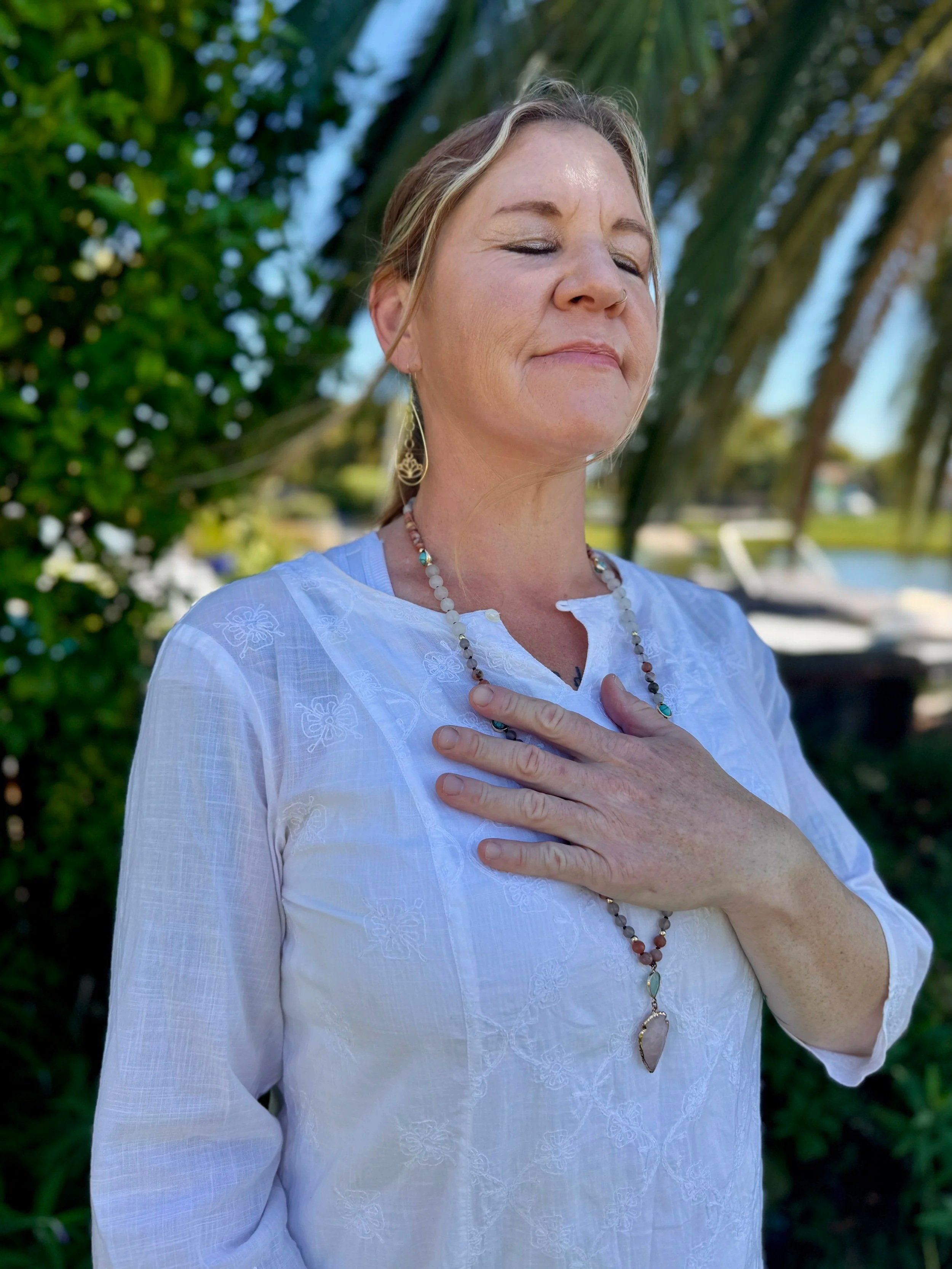 A woman in a white embroidered blouse with jewelry, standing outdoors with her eyes closed and hand on her chest, in front of greenery and trees.