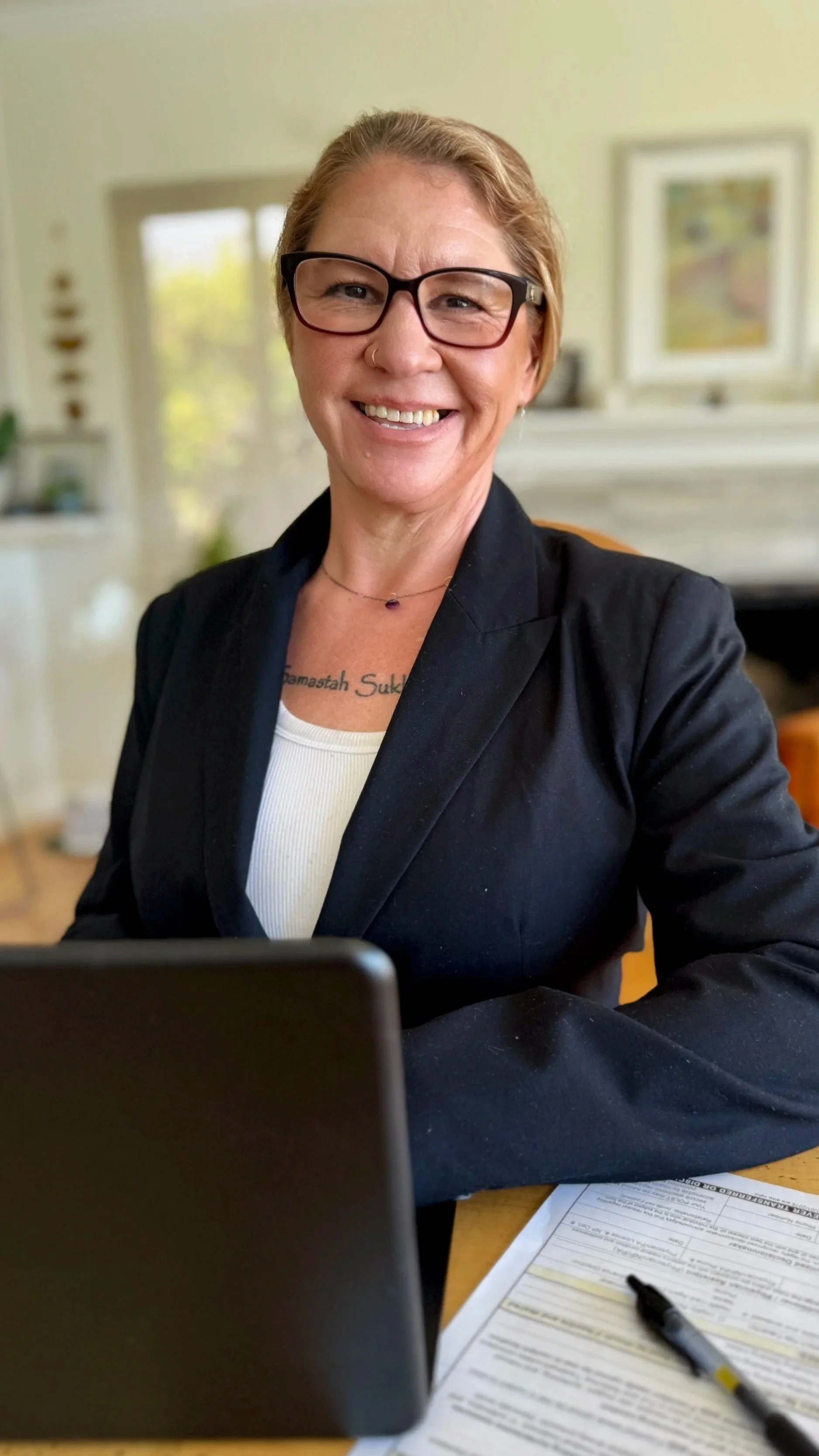 A woman with short blonde hair, glasses, and a nose ring smiling at the camera, wearing a black blazer and a white top, sitting at a desk with papers and a pen, in a well-lit home office.
