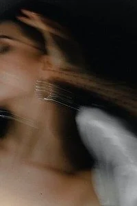 Blurred photo of a woman with long hair, wearing white, in low light.